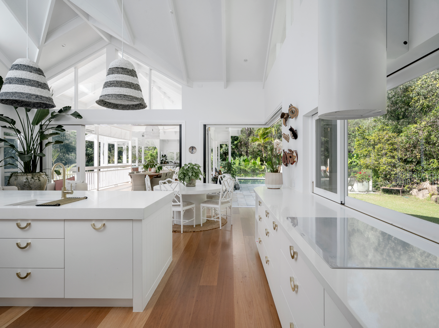 Bright white kitchen with large windows, white cabinetry, and a dining area with white chairs and a round table, overlooking a lush green garden.