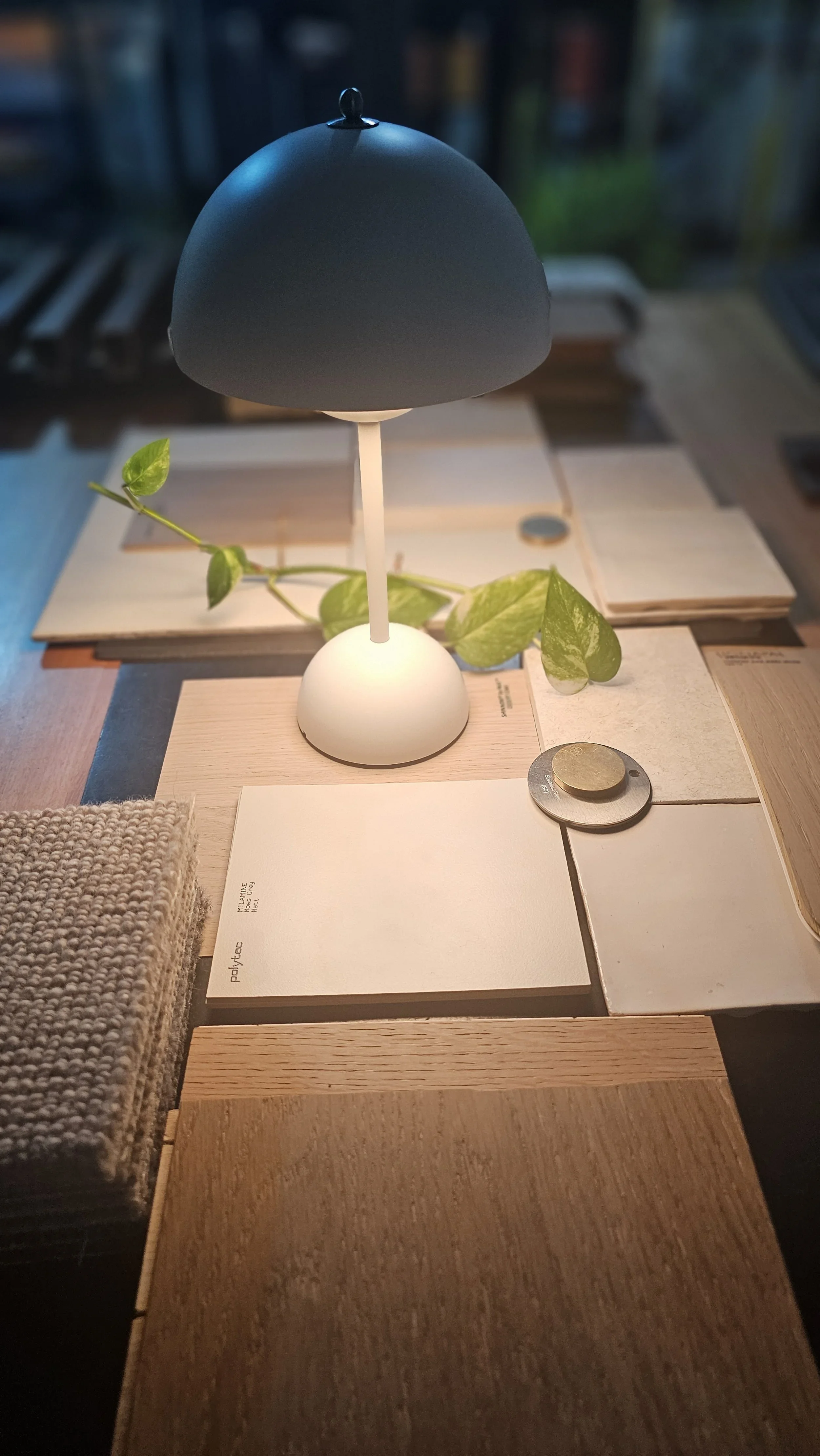 Close-up of a small modern table lamp with a black dome-shaped shade, a white base, and a white stem, placed on a wooden table surrounded by sample color and material swatches, along with a piece of green plant and some paper notes.