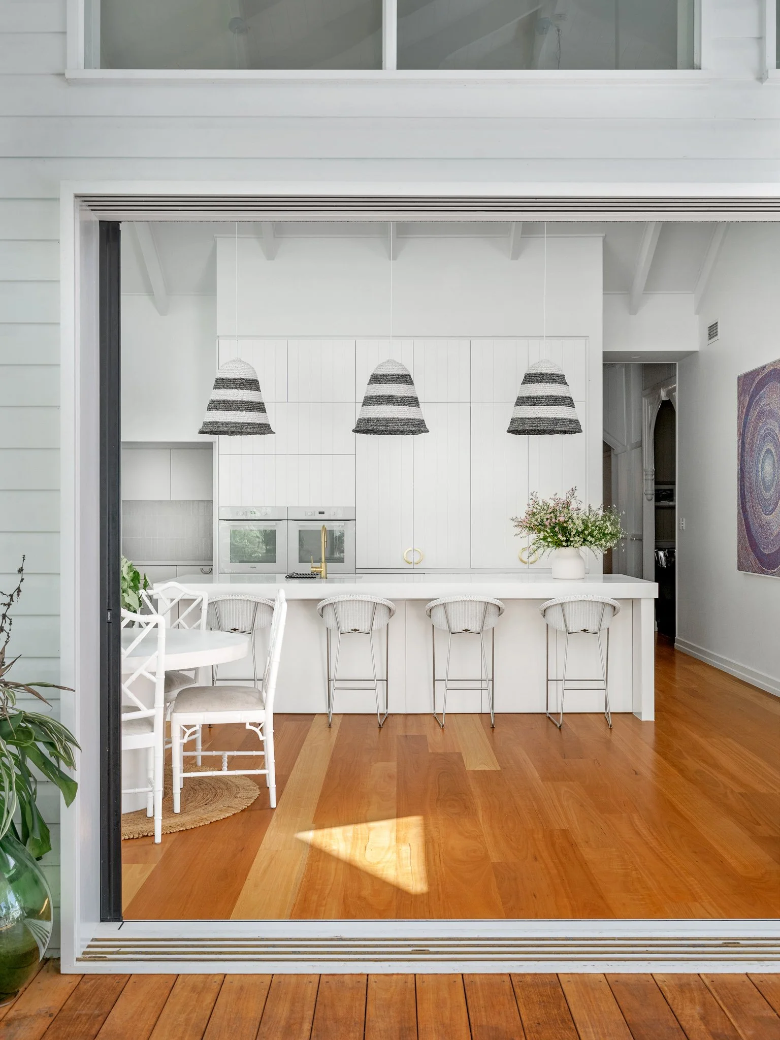 Open sliding glass door reveals a bright kitchen with white cabinets, a white kitchen island with bar stools, three woven pendant lights, and a bouquet of flowers on the countertop. Inside the kitchen, a built-in oven and a window are visible.