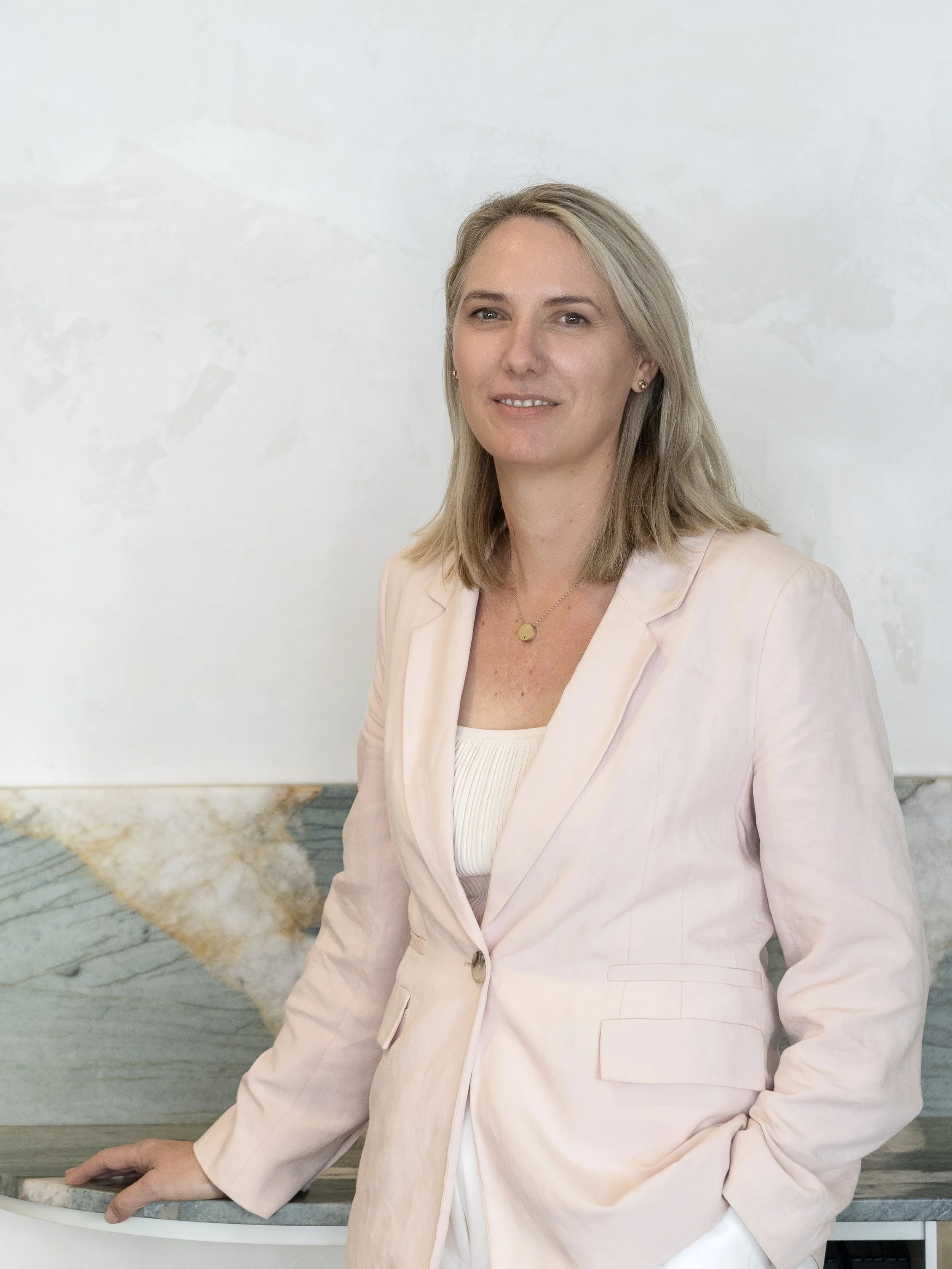 A woman with blonde hair wearing a light pink blazer and a necklace, sitting or leaning against a table with a marble surface, with a white background.