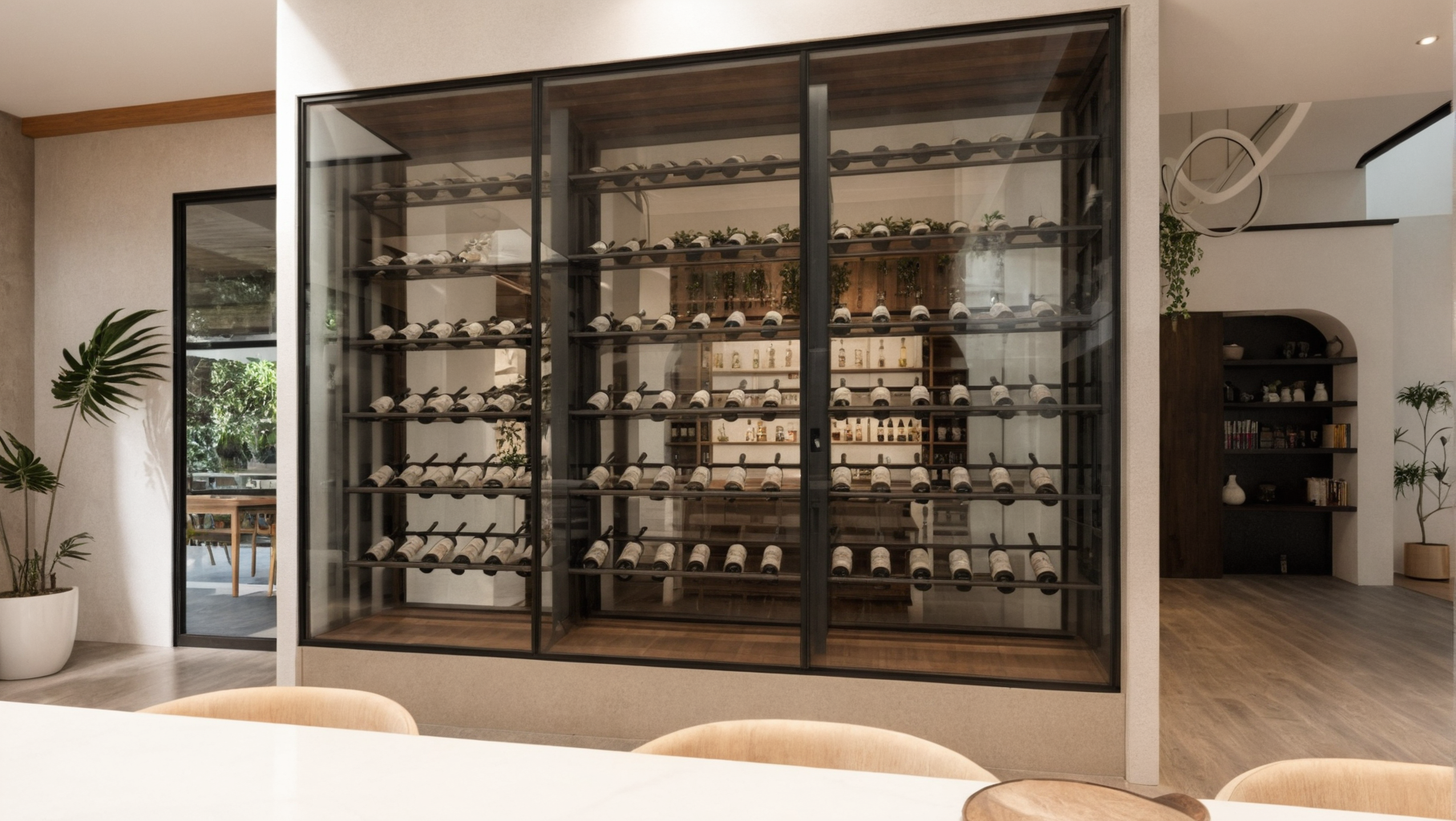 Modern wine cellar with glass door, containing multiple wine bottles stored on black metal racks. The room has a cozy, contemporary design with wooden accents, potted plants, and bookshelves visible in the background.