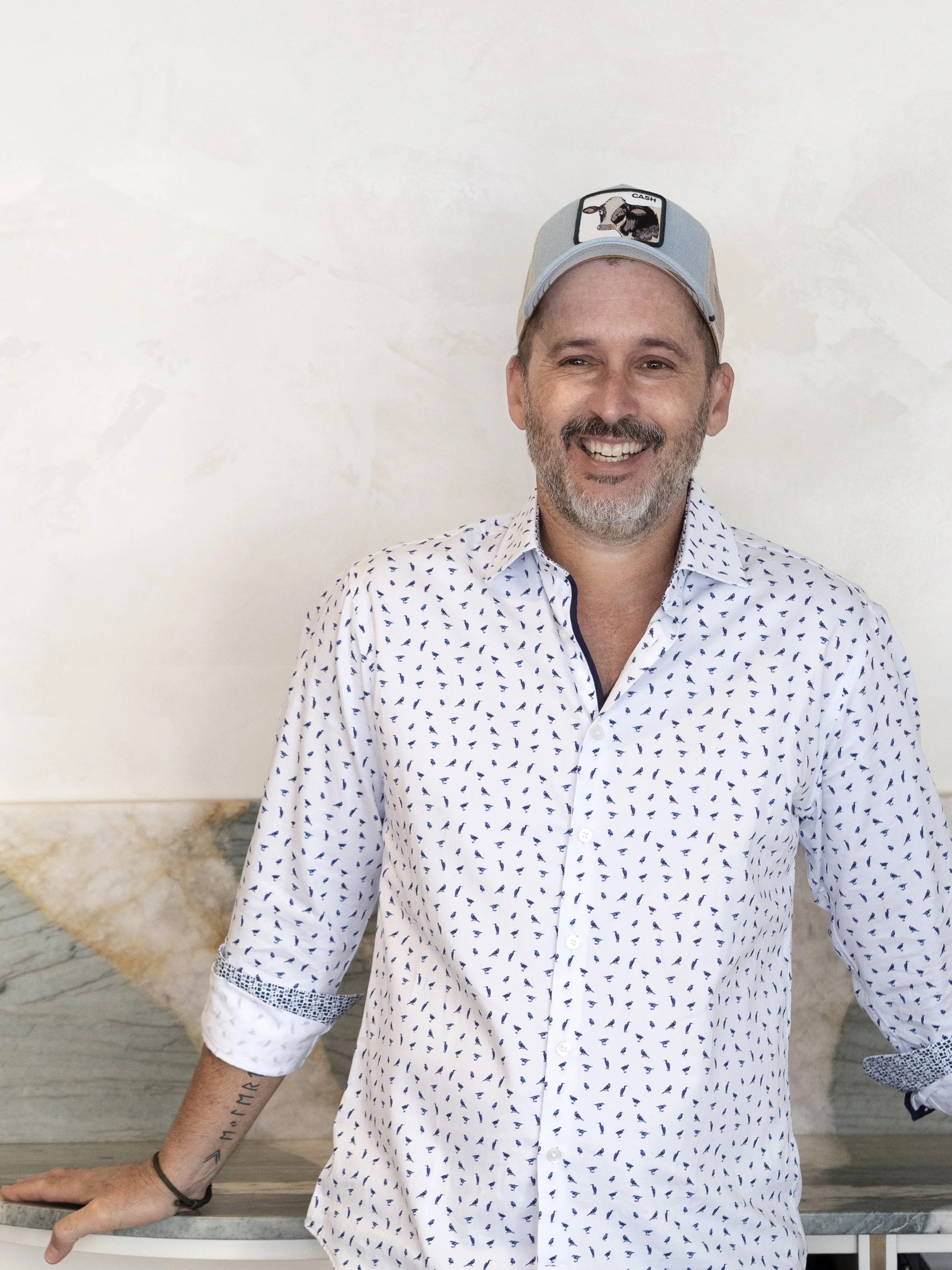 Smiling man wearing a white shirt with blue patterns, a baseball cap, and standing against a neutral background.