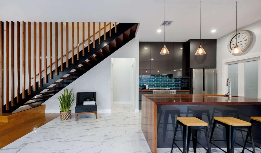 Modern kitchen and living space with marble flooring, black and wood staircase, kitchen island with stools, blue tile backsplash, stainless steel appliances, and wall clock.
