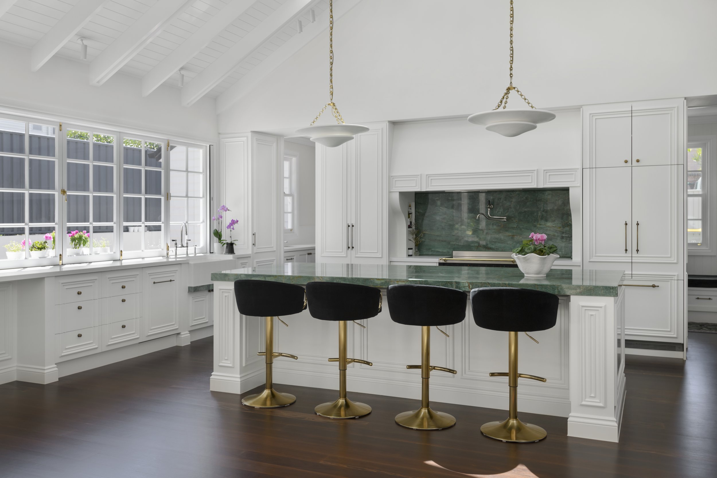 A modern kitchen with white cabinetry and a green marble island. Large windows with flower pots on the sill, dark wood flooring, and pendant lights hanging from a white beamed ceiling.