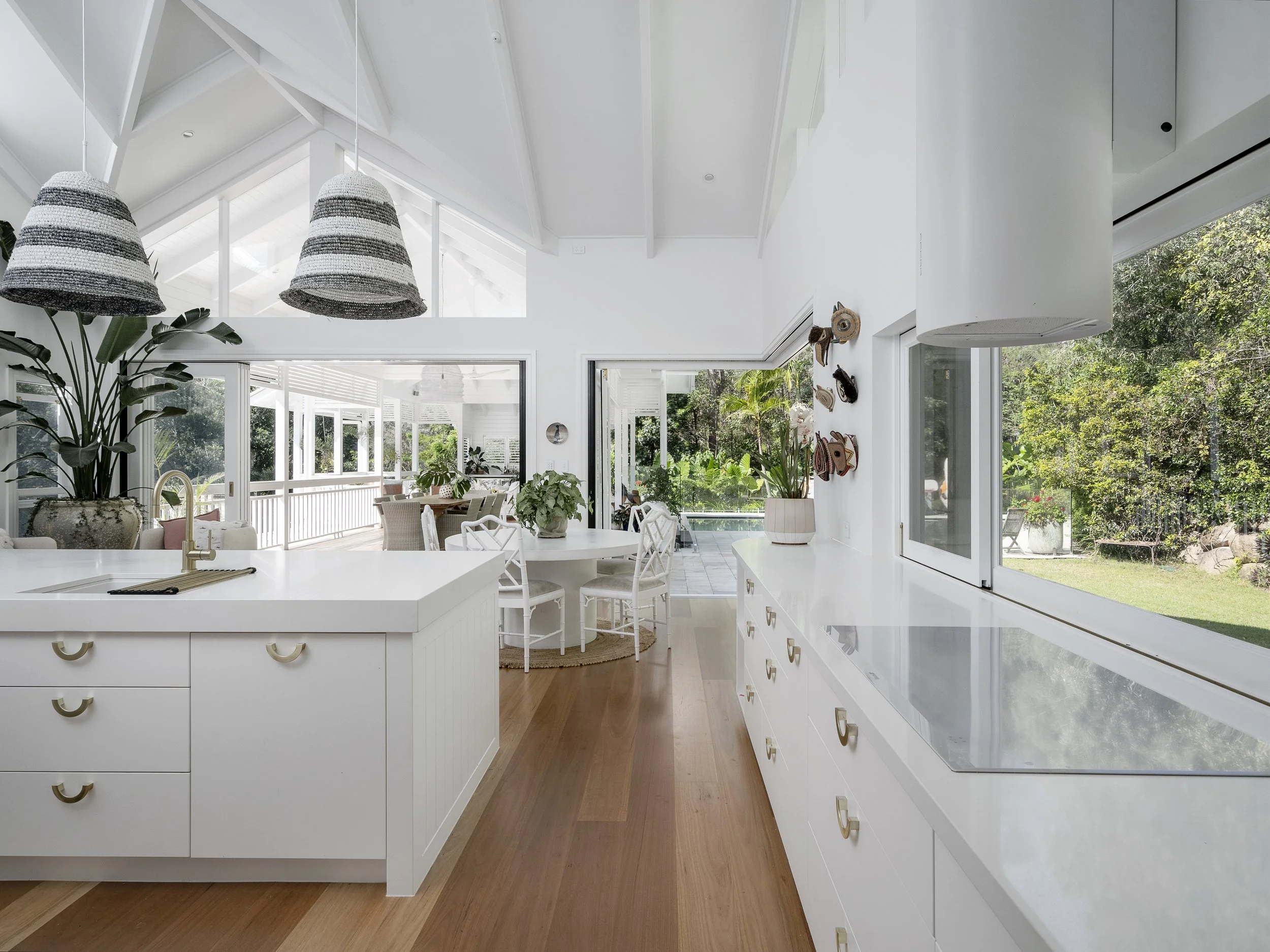 Bright modern kitchen and dining area with large windows, white cabinetry, a white island with a gold faucet, and potted plants, with a view of a lush green backyard.