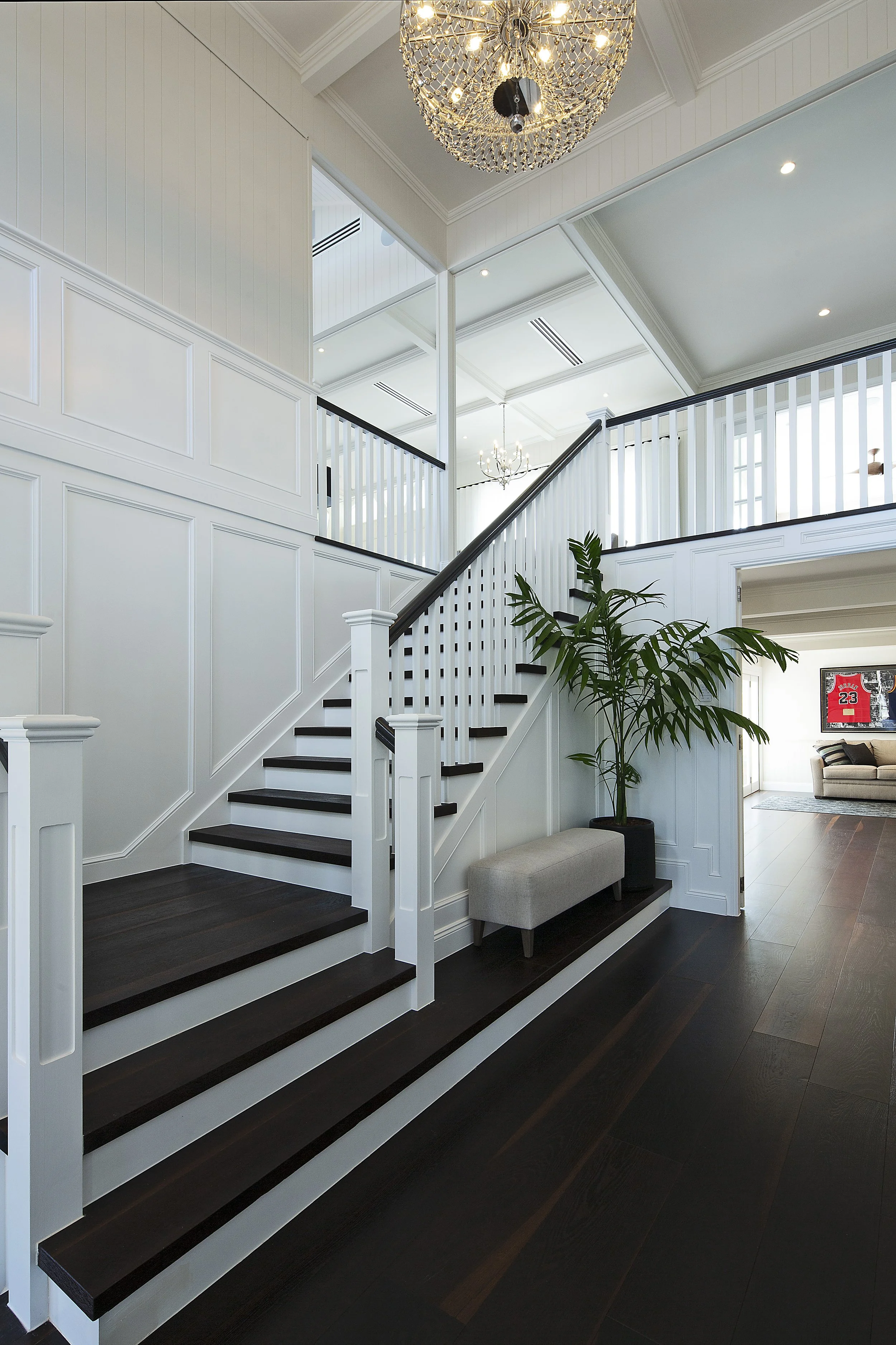 A modern staircase with dark wooden steps and white risers, leading to an upper floor with white railings and paneling. There is a large green plant at the bottom of the stairs, and a beige bench underneath the staircase. The ceiling features crown molding, with two hanging chandeliers and exposed ceiling lights, in a spacious, well-lit home interior.