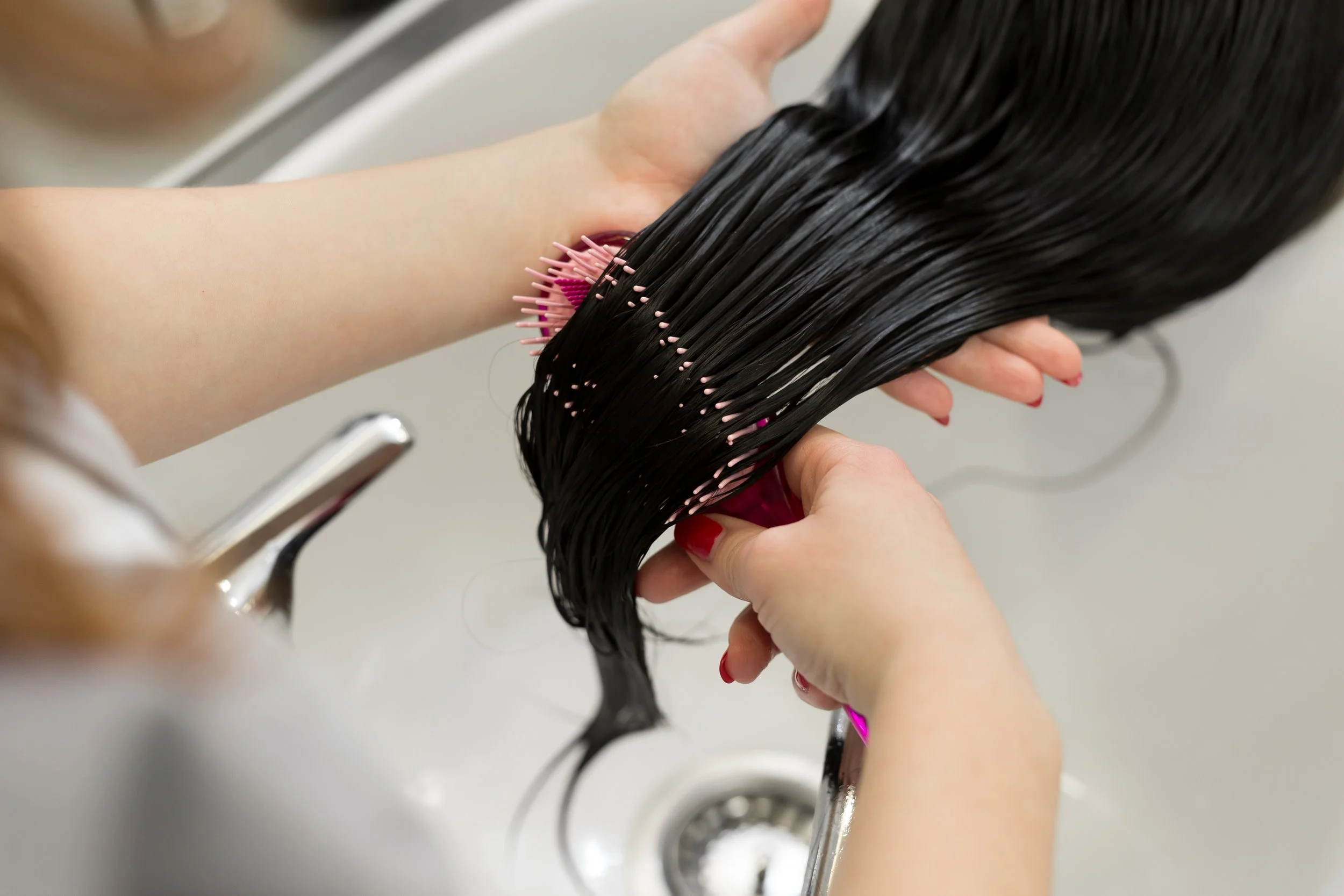 Person washing long, straight, black hair in a sink with a pink hairbrush.