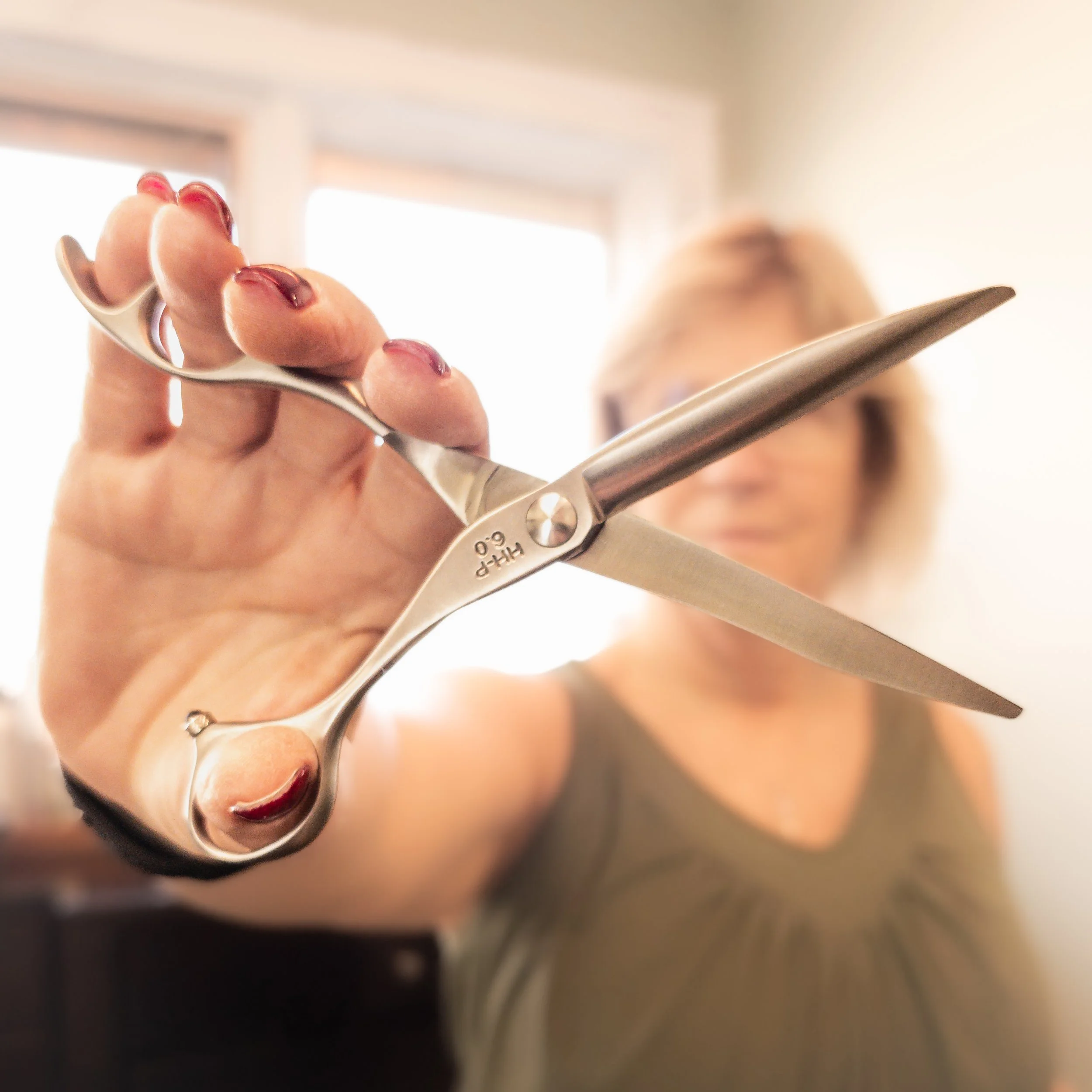 A woman holds a pair of scissors close to the camera, with the blades open, in a brightly lit room.