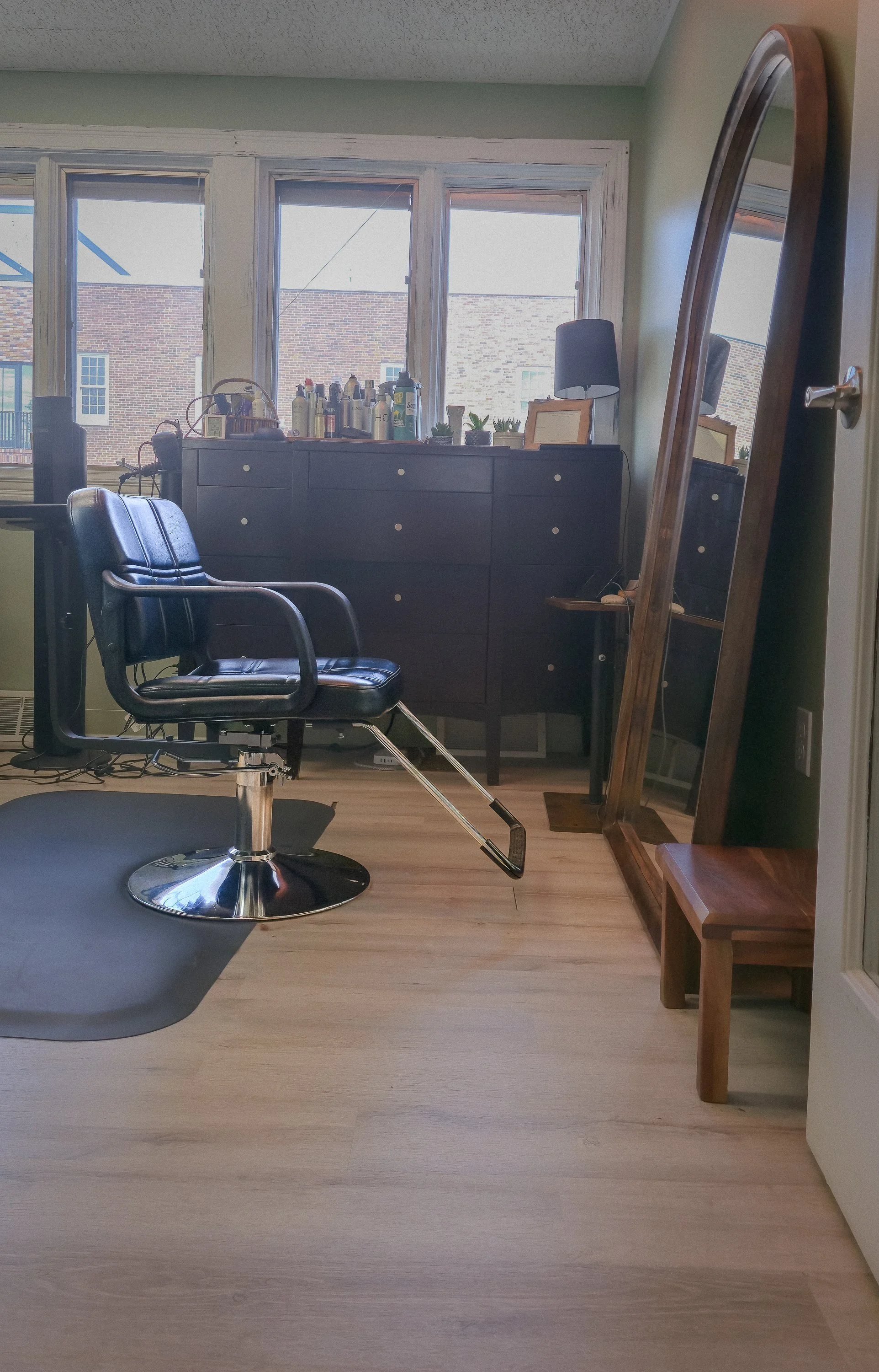 Studio interior with a black salon chair, a large standing mirror, a wooden sideboard with bottles and small plants on top, and a window with a view of brick buildings outside.
