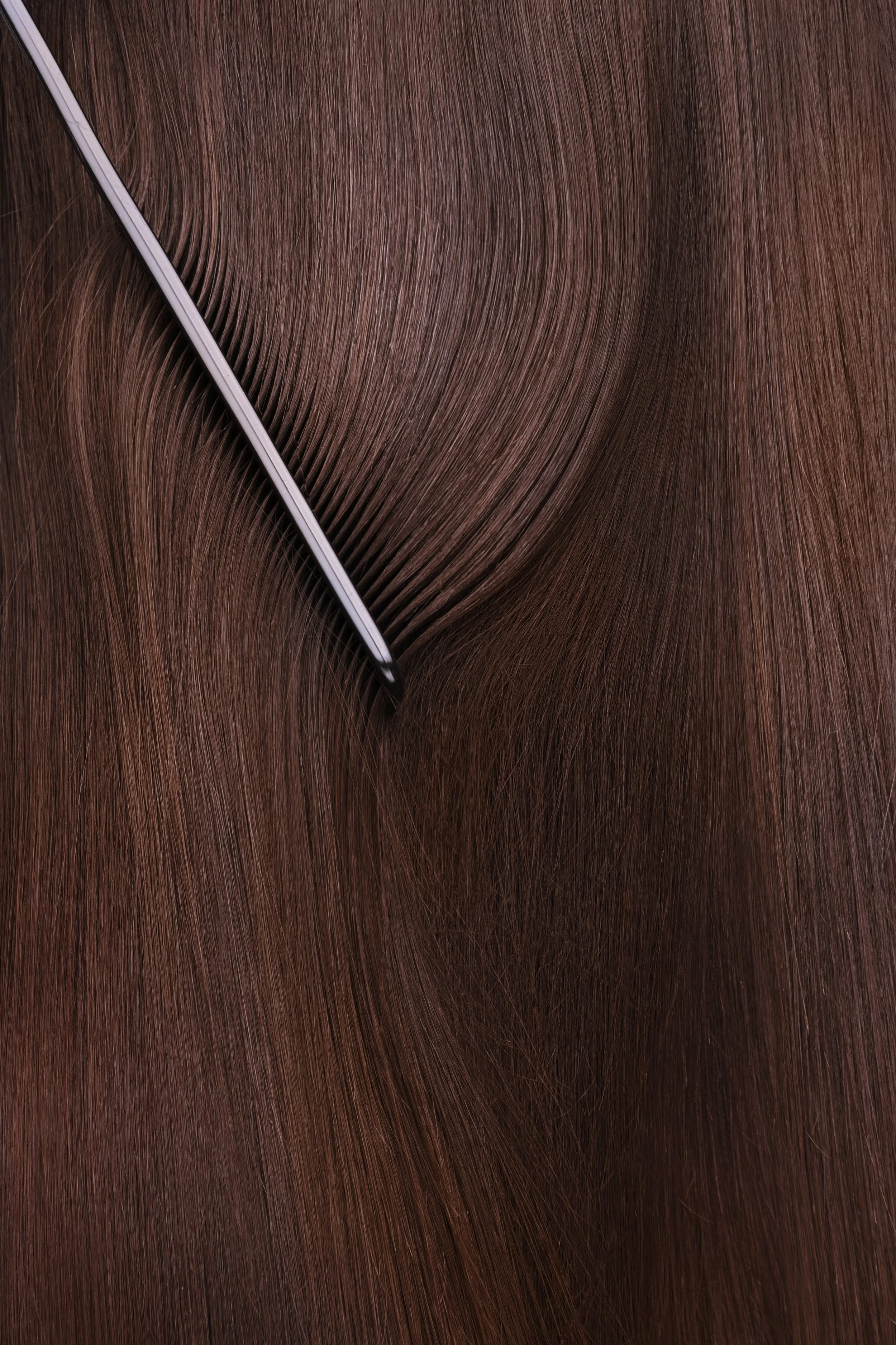 Close-up of a lock of smooth, brown hair with a comb pulling the top layer to the side.