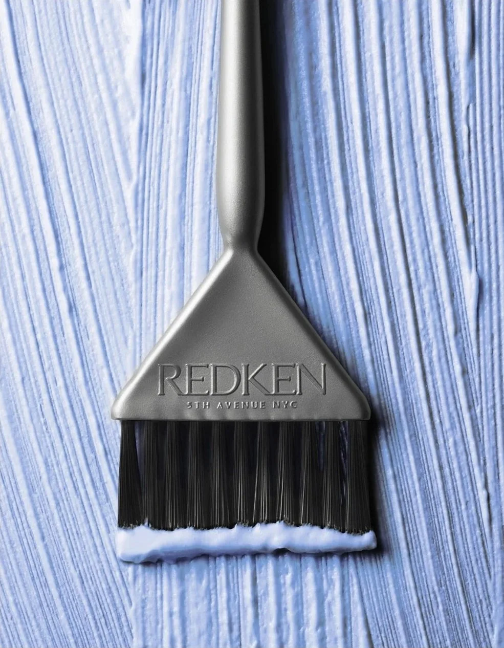 A hair dye brush with a gray handle engraved with 'REDKEN 5TH AVENUE NYC', placed on a surface covered with light blue textured material.