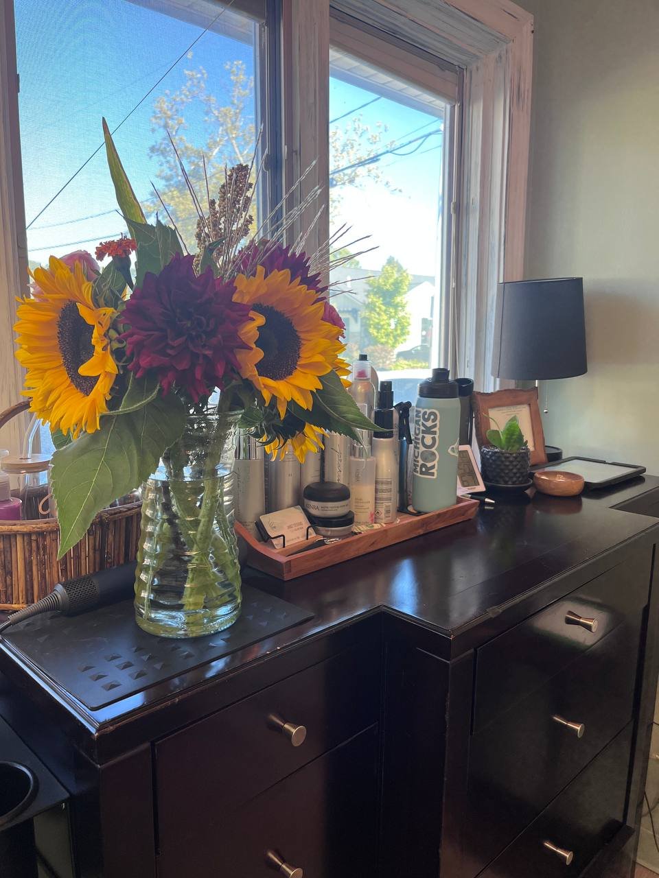 A black dresser with a vase of sunflower and purple flowers, and various personal items on a tray and surface near a window with trees visible outside.