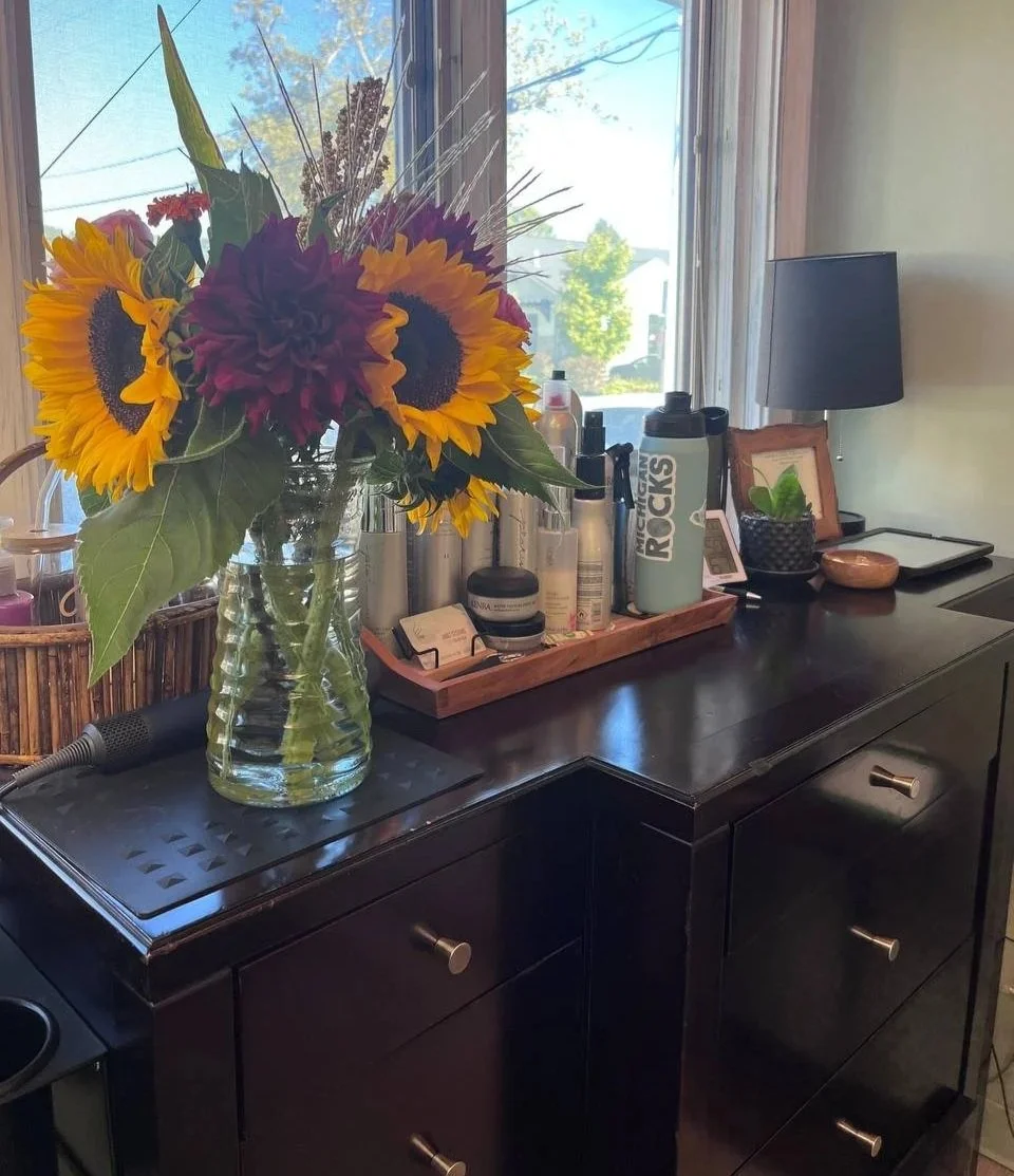 A black sideboard with a glass vase of sunflower, red flower, and purple flower on top next to a window, with various skincare and drink bottles, a tissue box, framed photo, small lamp, and electronic devices.