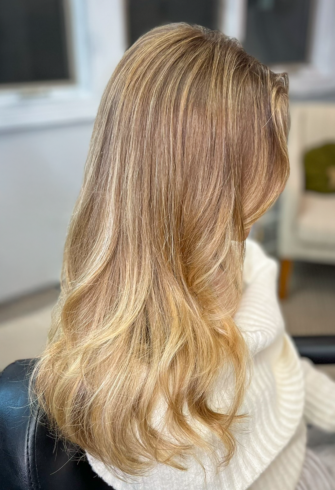 A woman with long, wavy, light brown hair sitting indoors, wearing a white sweater.