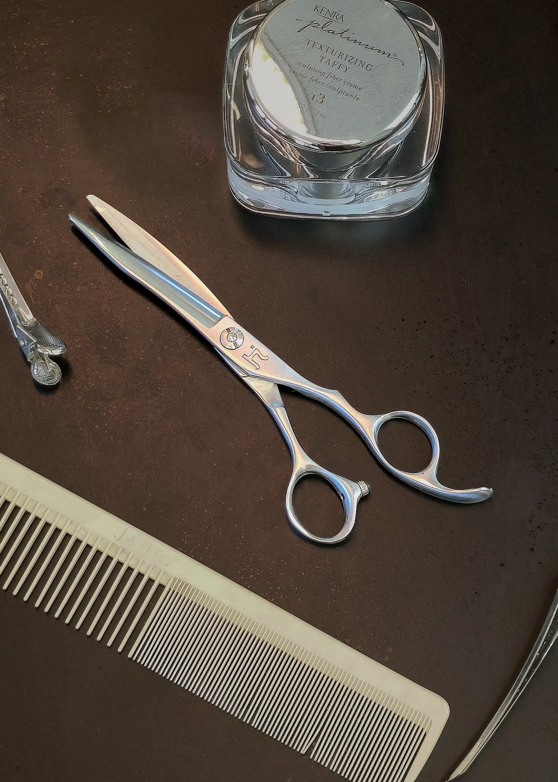 A pair of silver Hattori Hanzo scissors, a white comb, and a container of Kenra platinum texturizing spray are on a dark surface.