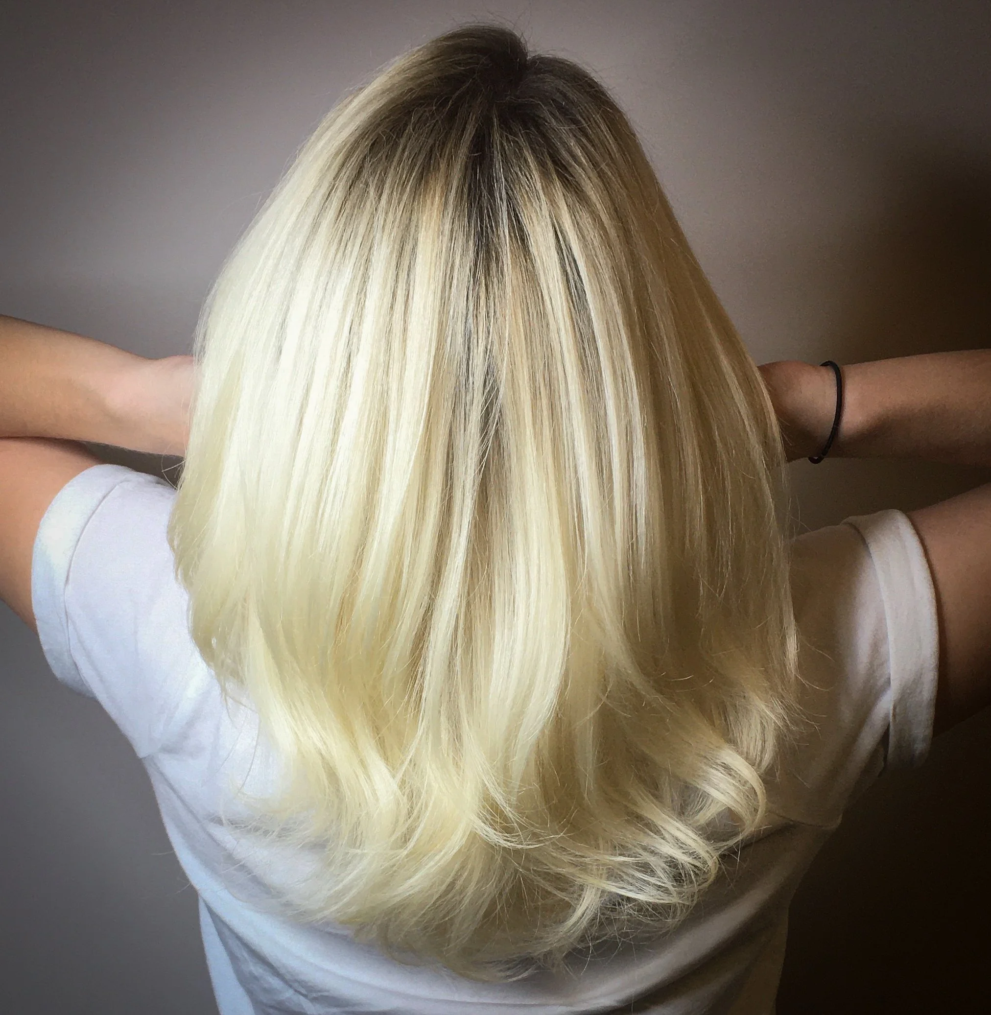 Back of a woman with platinum blonde, wavy hair, wearing a white shirt and a black hair tie, with arms raised and hands behind her head.