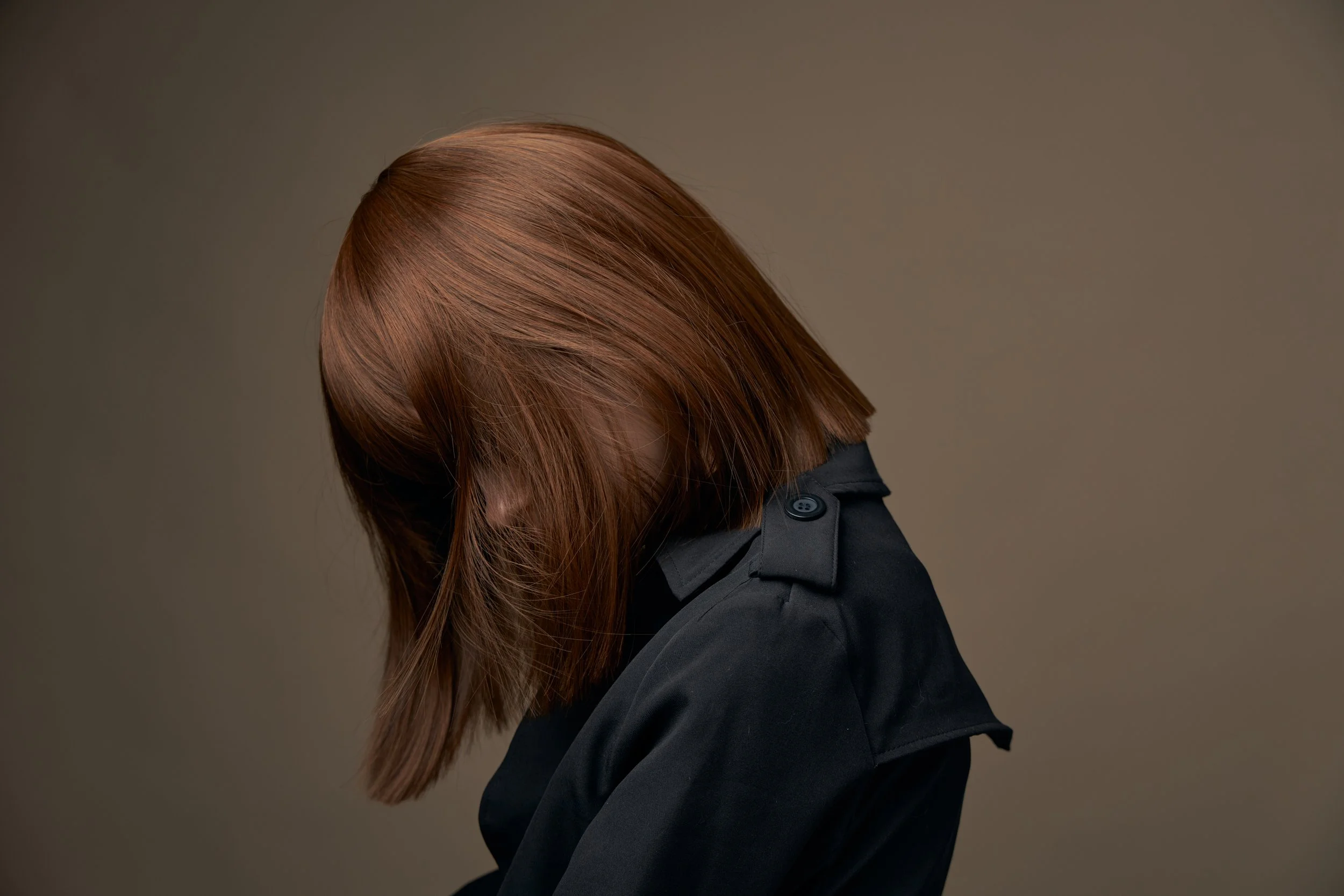 A person with shoulder-length reddish-brown hair wearing a black coat, facing downward, with an out-of-focus neutral background.