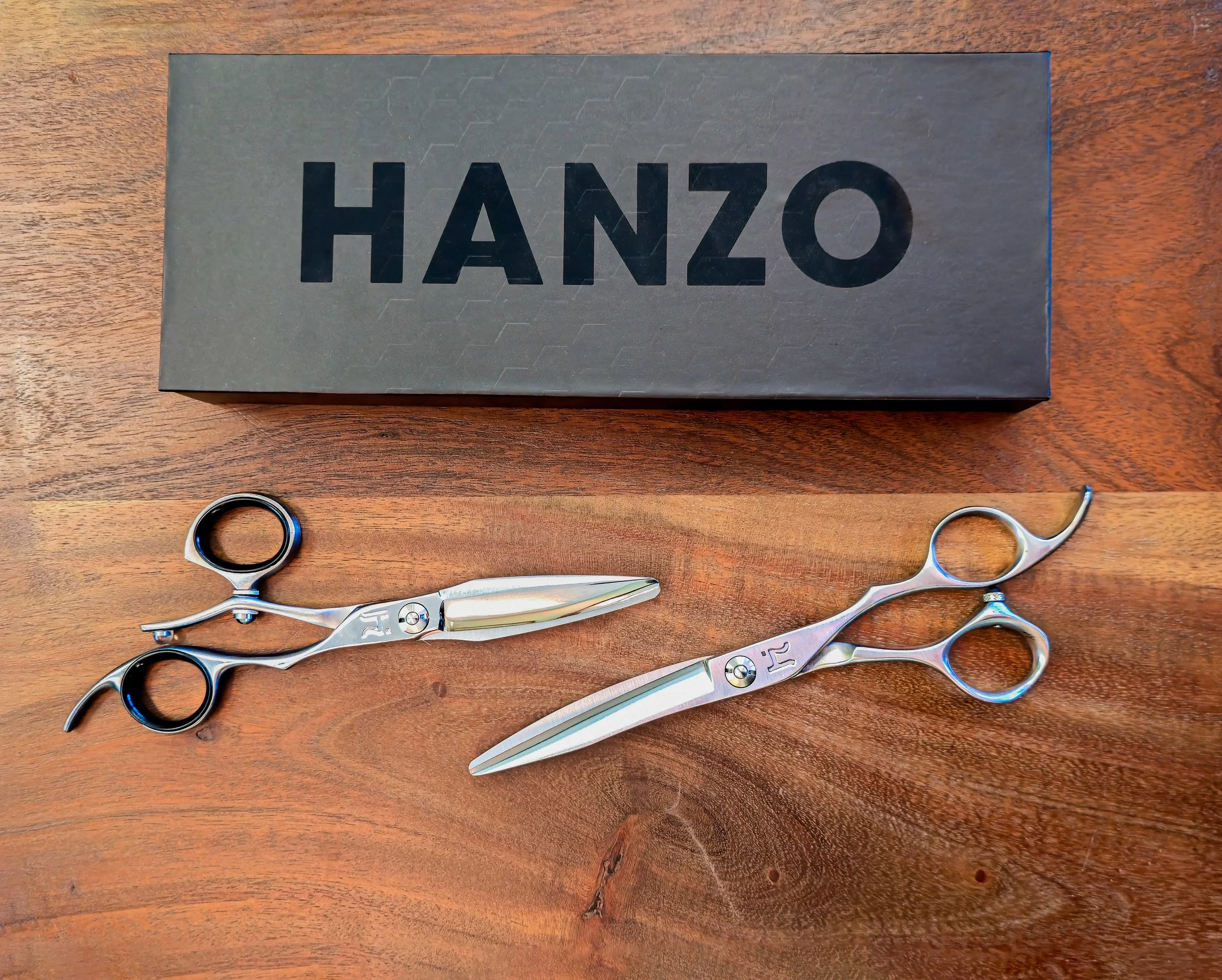 A black box with the word "HANZO" in large letters, and two barber scissors with silver handles and blades, placed on a wooden surface.