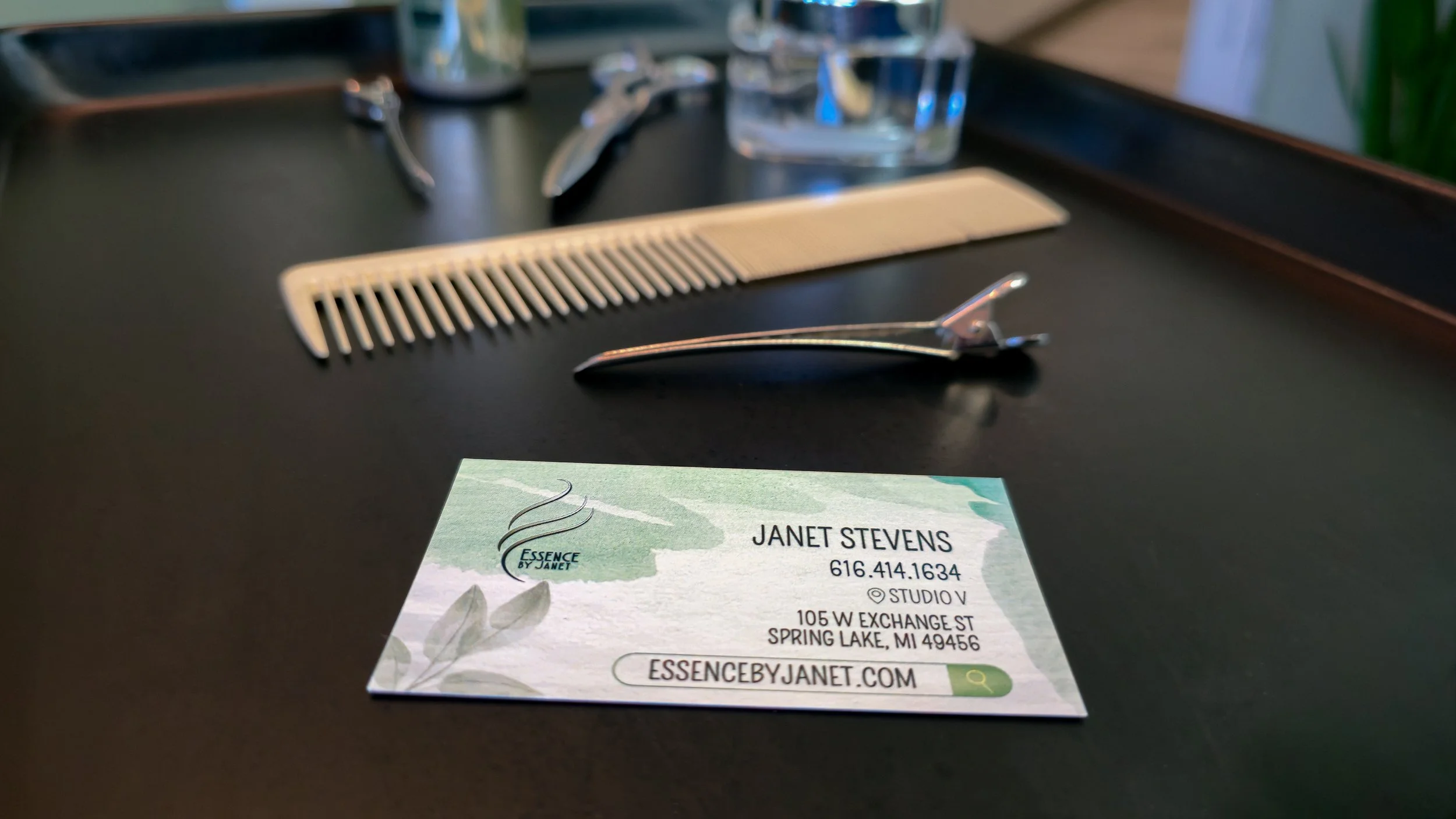 Business card for Janet Stevens from Essence by Janet on a dark tabletop, with hairdressing tools including a white comb and hair clips, and a Kenra styling bottle in the background.