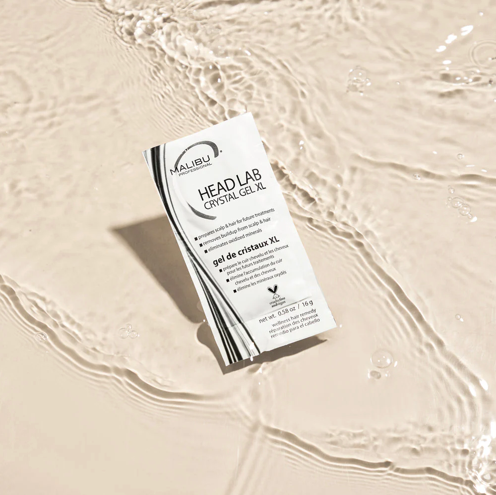 A box of Malibu Professional Head Lab Crystal Gel XL hair treatment lying on beige sandy surface with water ripples.