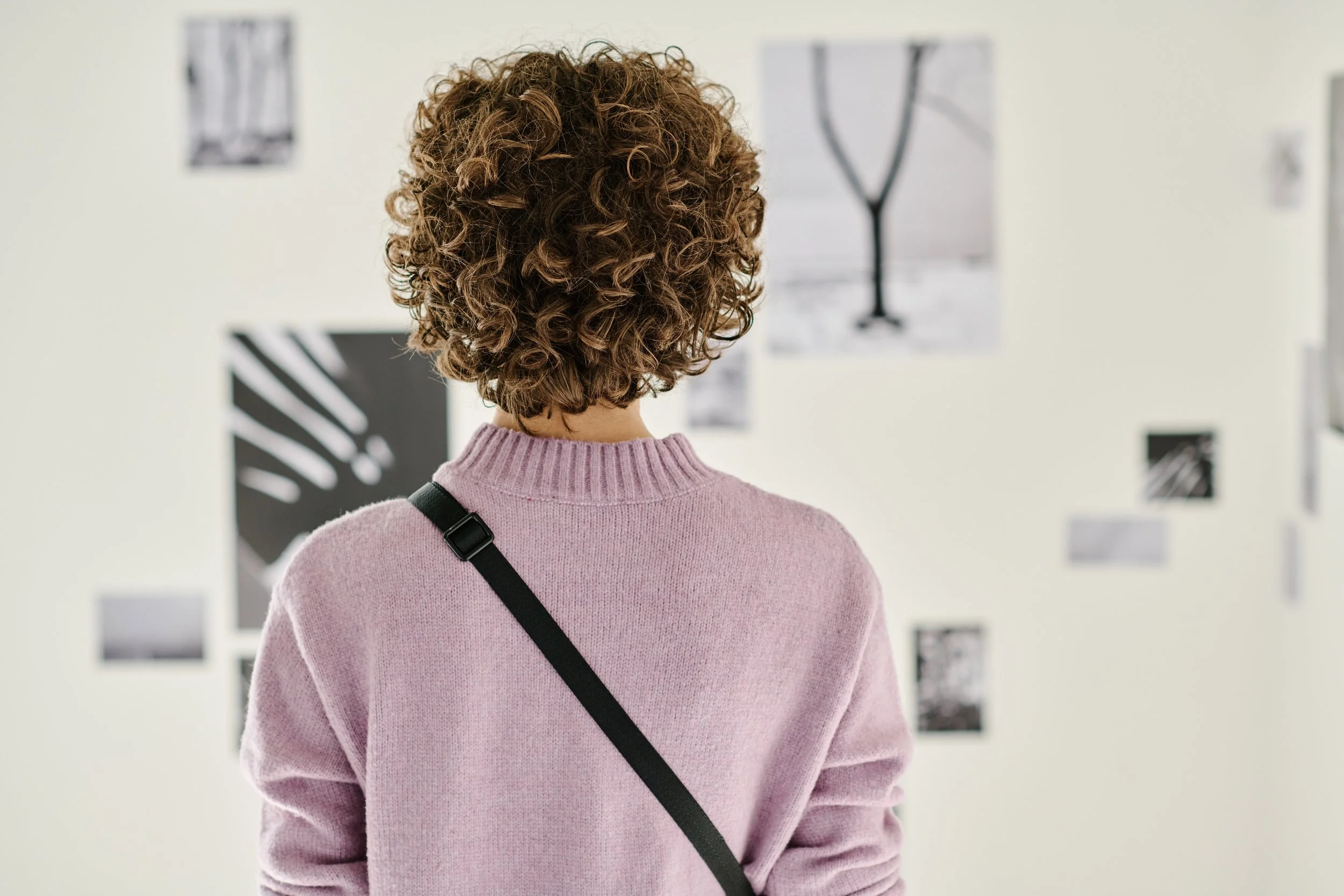 Person with curly hair wearing a pink sweater viewing black and white photographs on a wall.