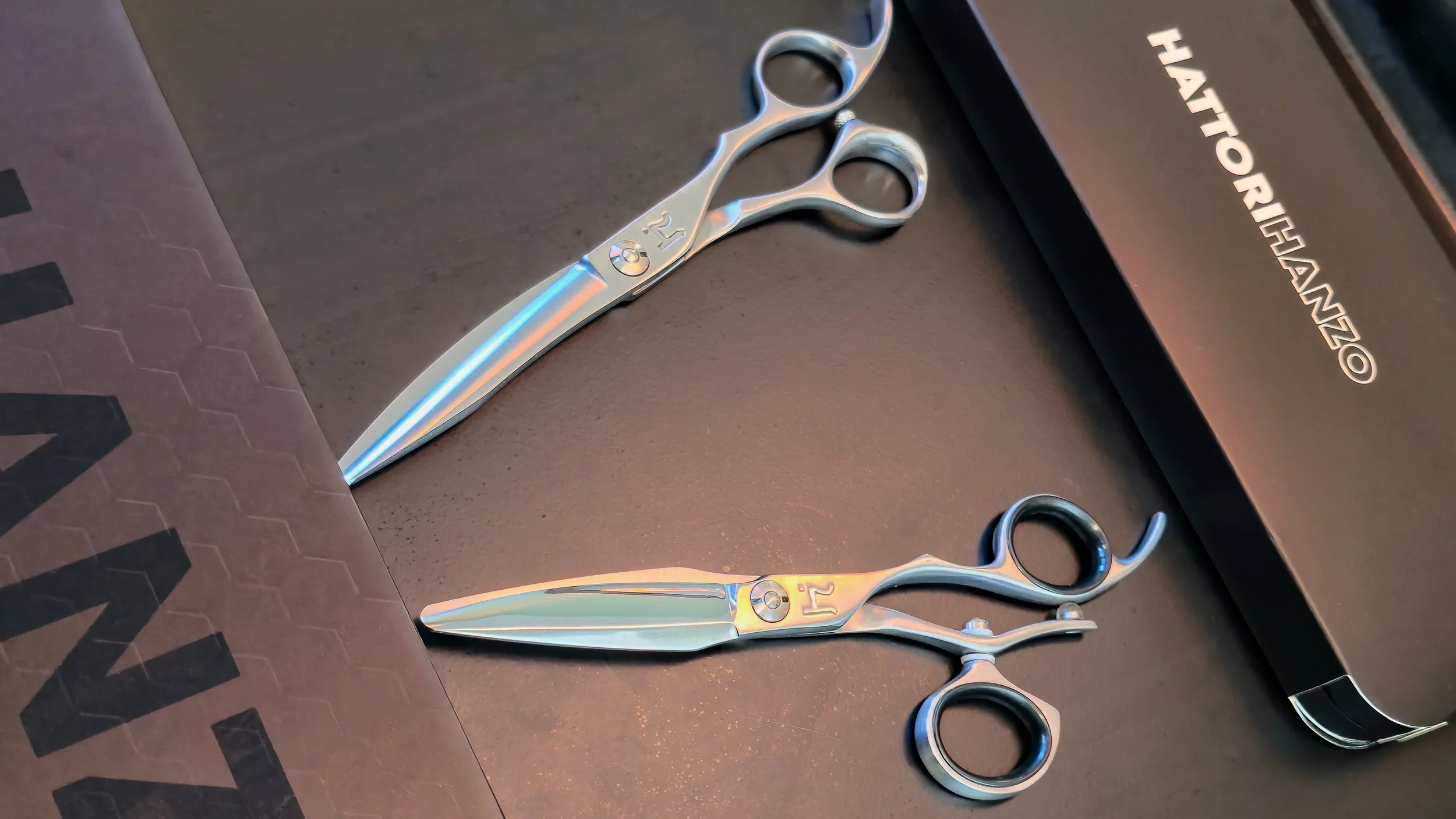 Two pairs of professional scissors, placed on a black surface near a box with the words 'HATTORI HAZO' on it.