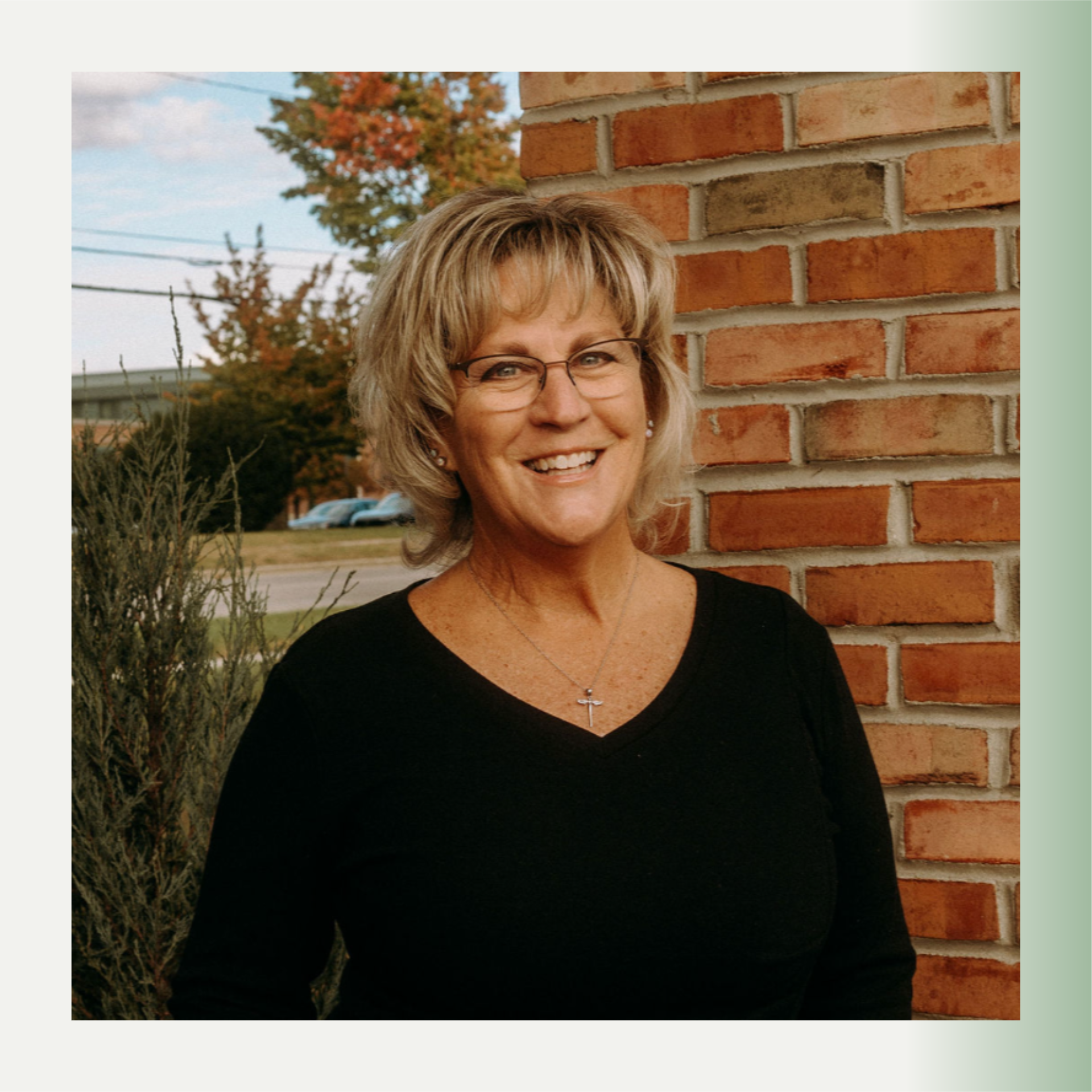 A smiling middle-aged woman, Janet, with blonde hair, glasses, and a cross necklace, standing next to a brick wall outdoors during fall.