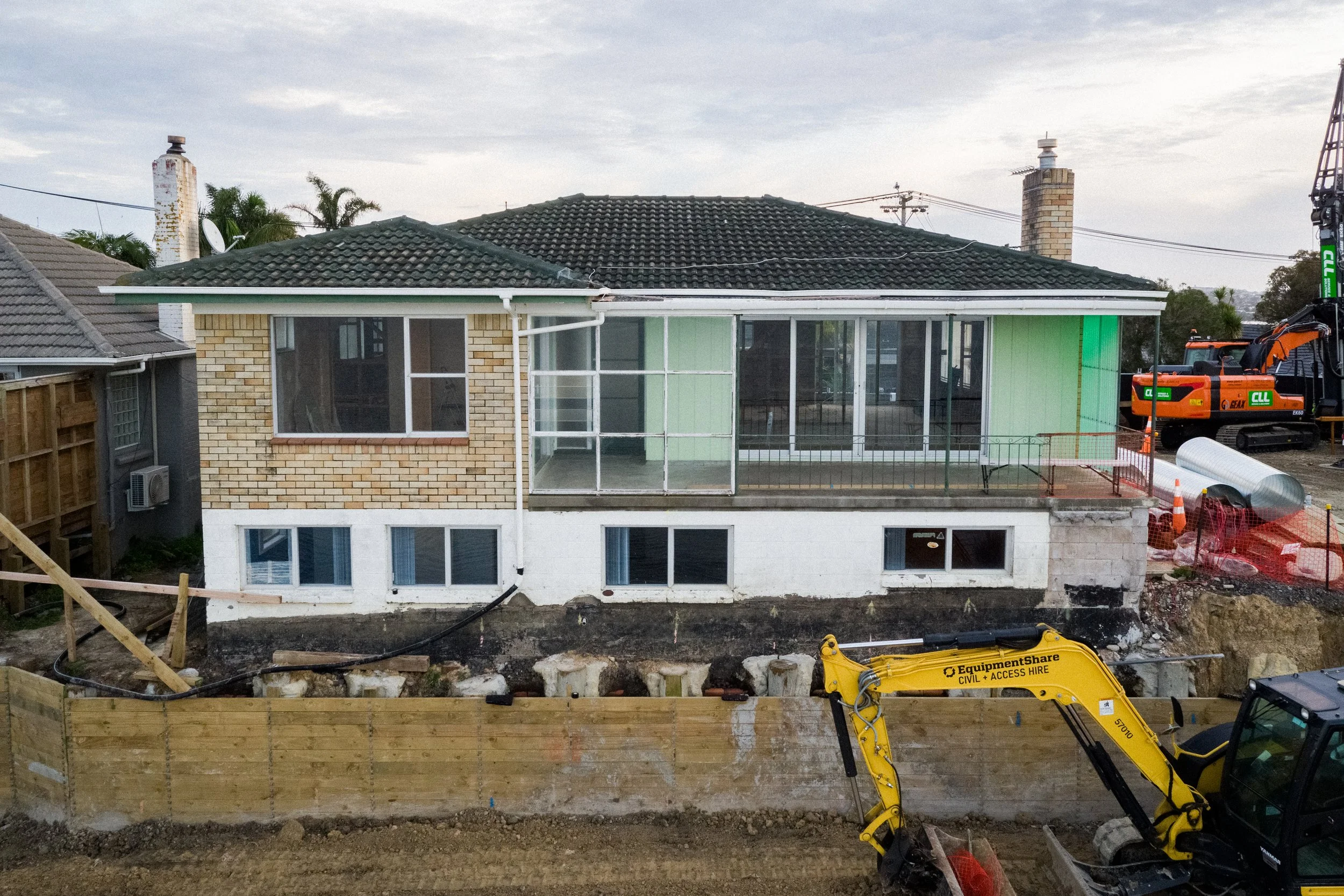 Construction site in a residential area with a house being renovated or built, construction equipment like an excavator, and a partially completed wooden fence.