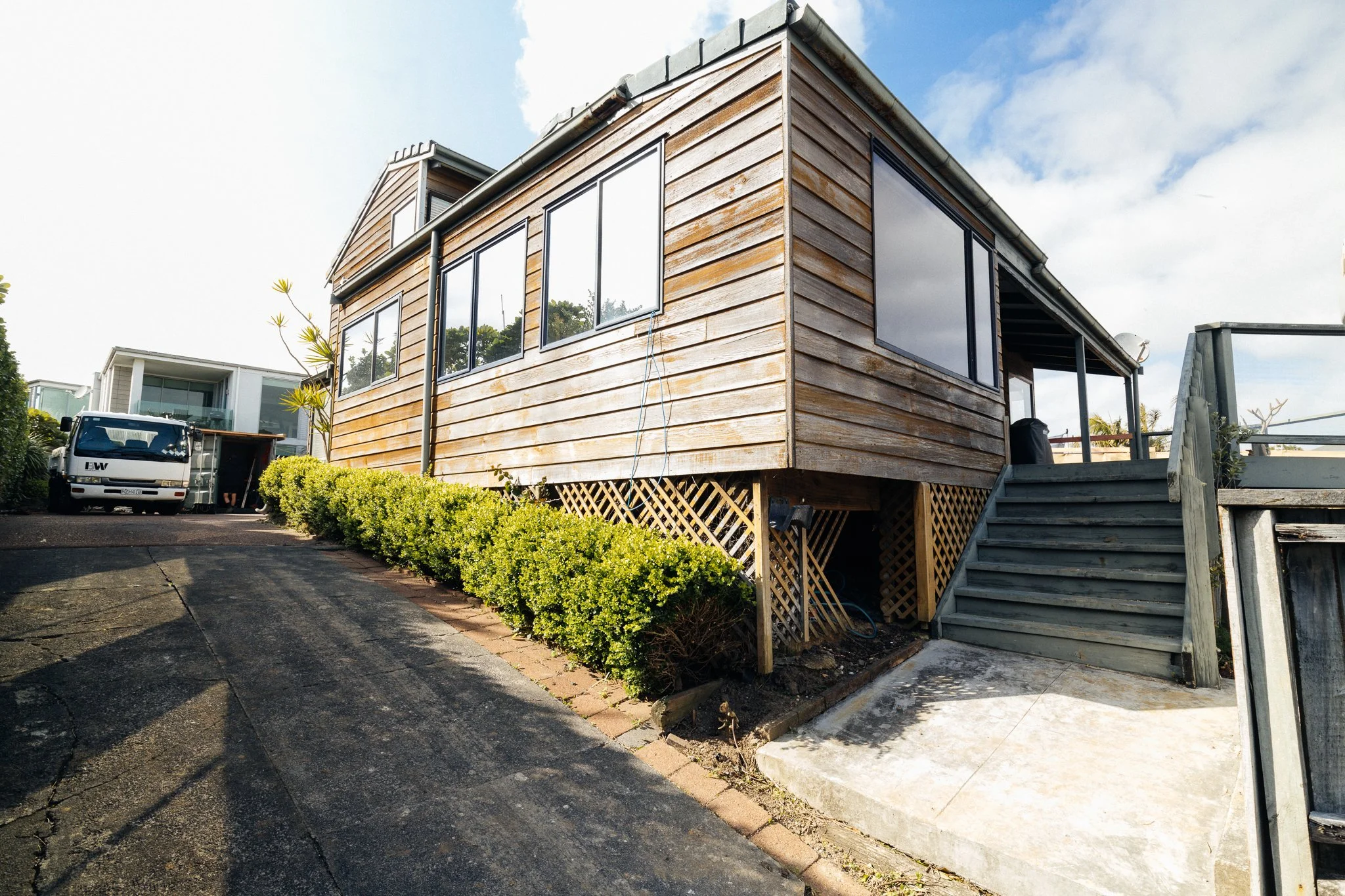 Wooden house with large windows on a raised foundation, stairs leading to the porch, and surrounded by greenery under a partly cloudy sky.