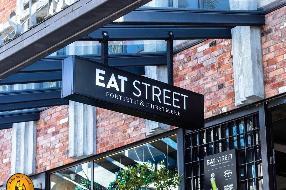Sign for Eat Street restaurant on Fortieth & Hurstmere street in an urban area with brick building and glass storefront.