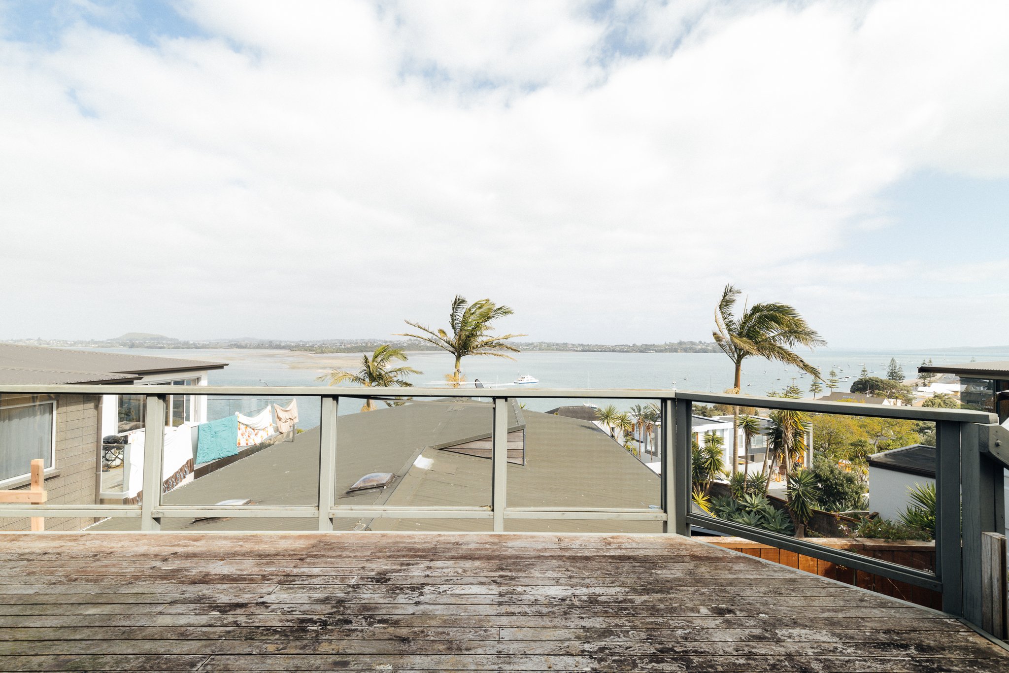 Balcony view showing palm trees, rooftops, and a body of water with boats, under a cloudy sky.