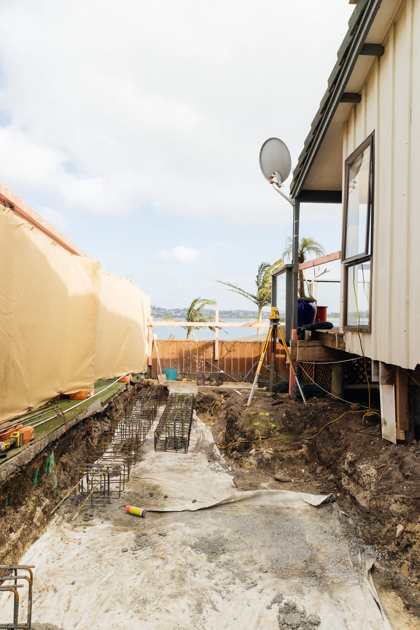 Construction site with exposed ground, rebar, and foundation work in progress, near a building with a satellite dish and open window, overlooking a scenic landscape with palm trees and water.