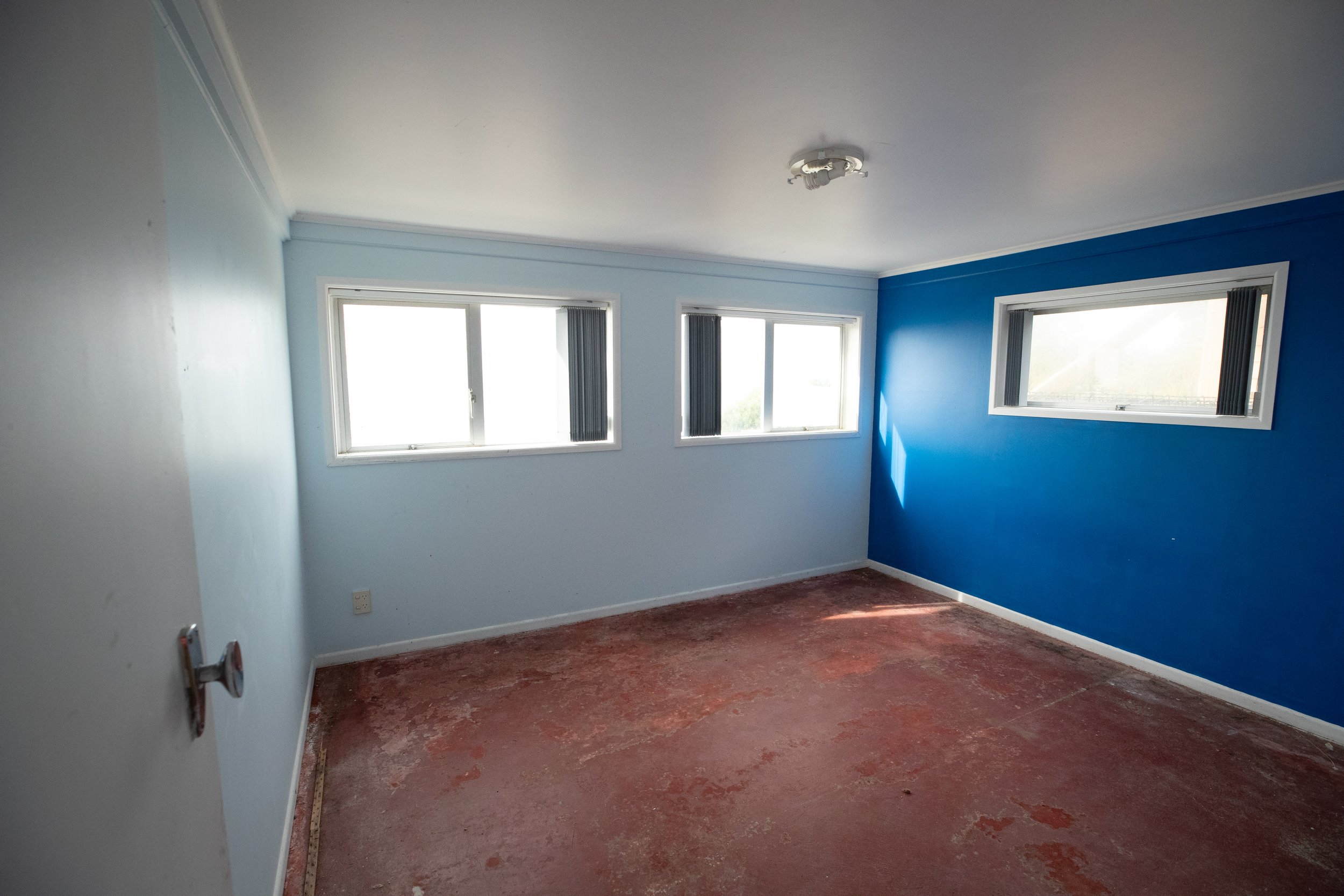 Empty room with light blue and dark blue walls, three windows, and unfinished reddish-brown floor.