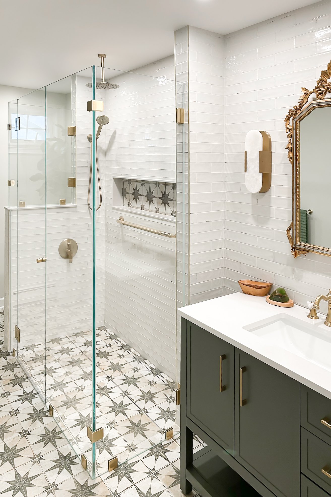 Modern bathroom with glass shower, white subway tiles, decorative floor tiles, dark green vanity with brass fixtures, ornate mirror, and round wall sconce.