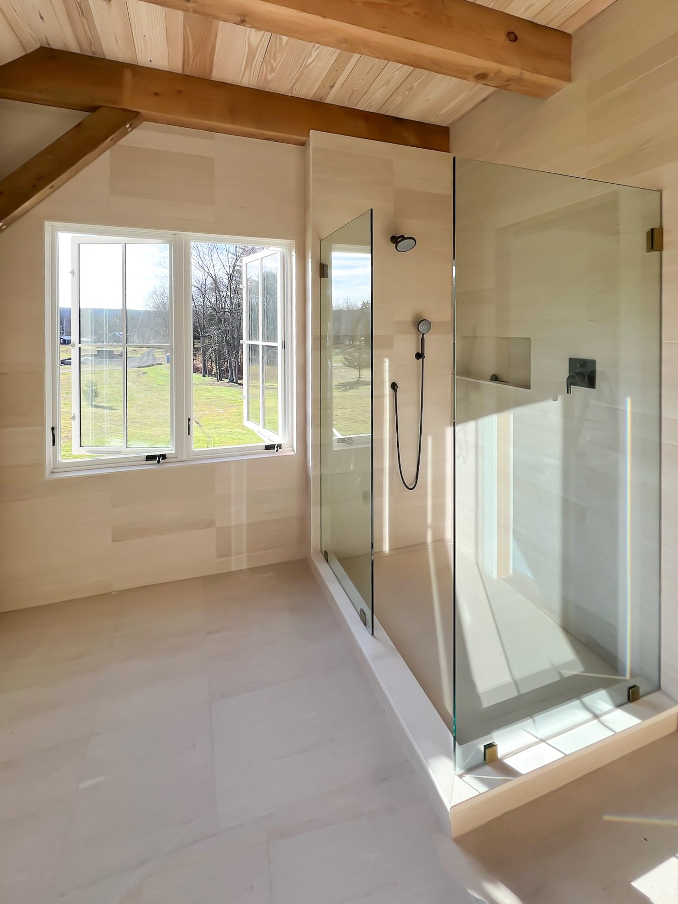 Modern bathroom with large glass shower enclosure, beige tiles, wood ceiling beams, and a large window overlooking grassy outdoor area.