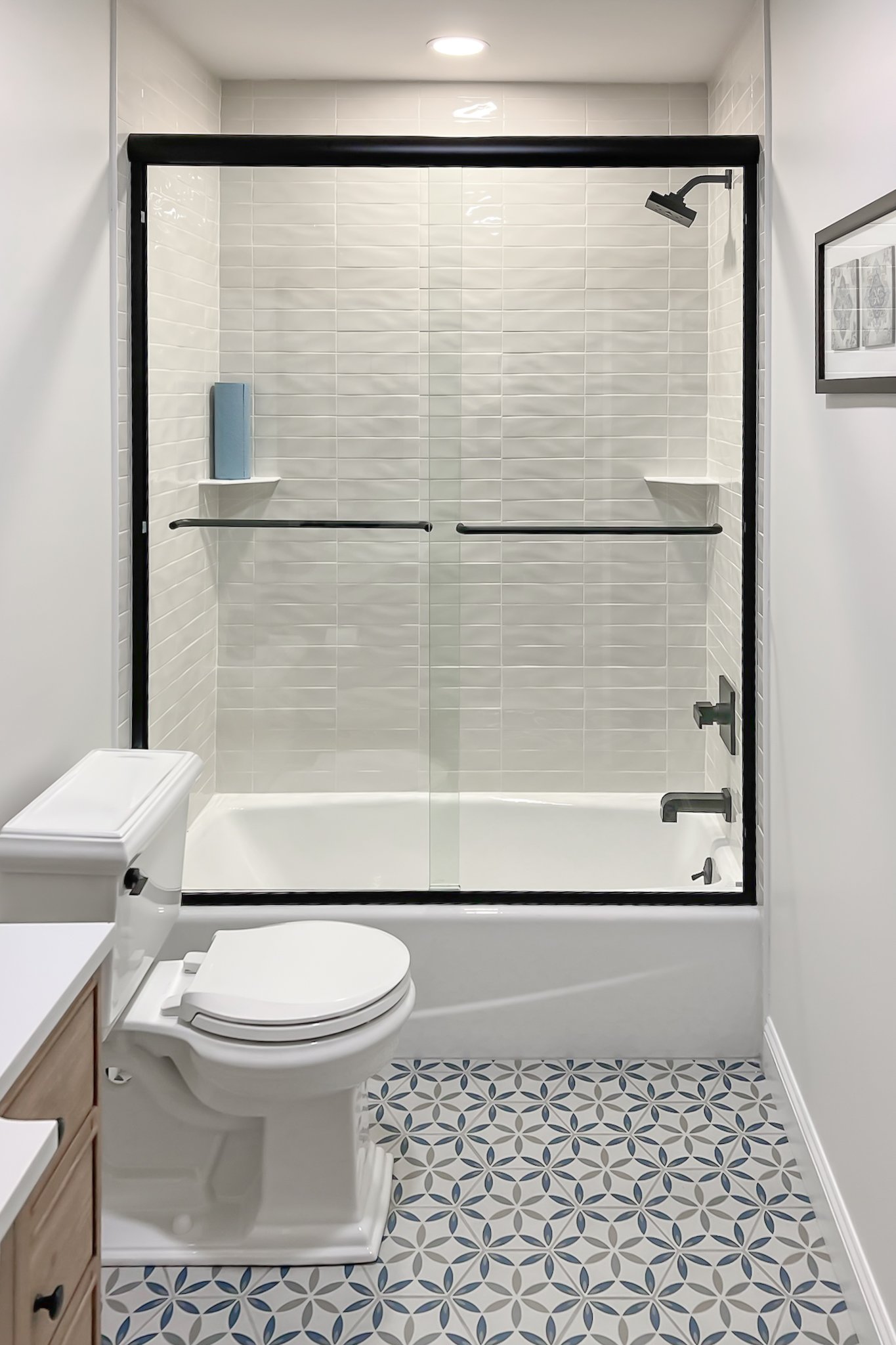 Modern bathroom featuring a toilet, patterned tile floor, and a shower-tub combo with glass doors, white tiles, and black fixtures.