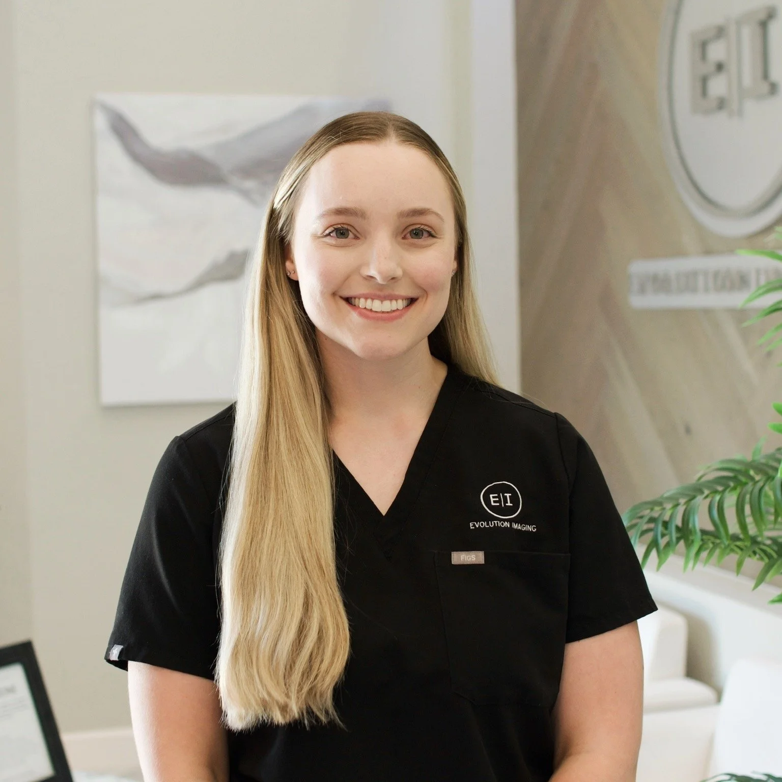 A young woman with long blonde hair and a black shirt with a logo reading 'E I' and 'Evolution Imaging,' smiling in an office setting with abstract wall art and a plant in the background.