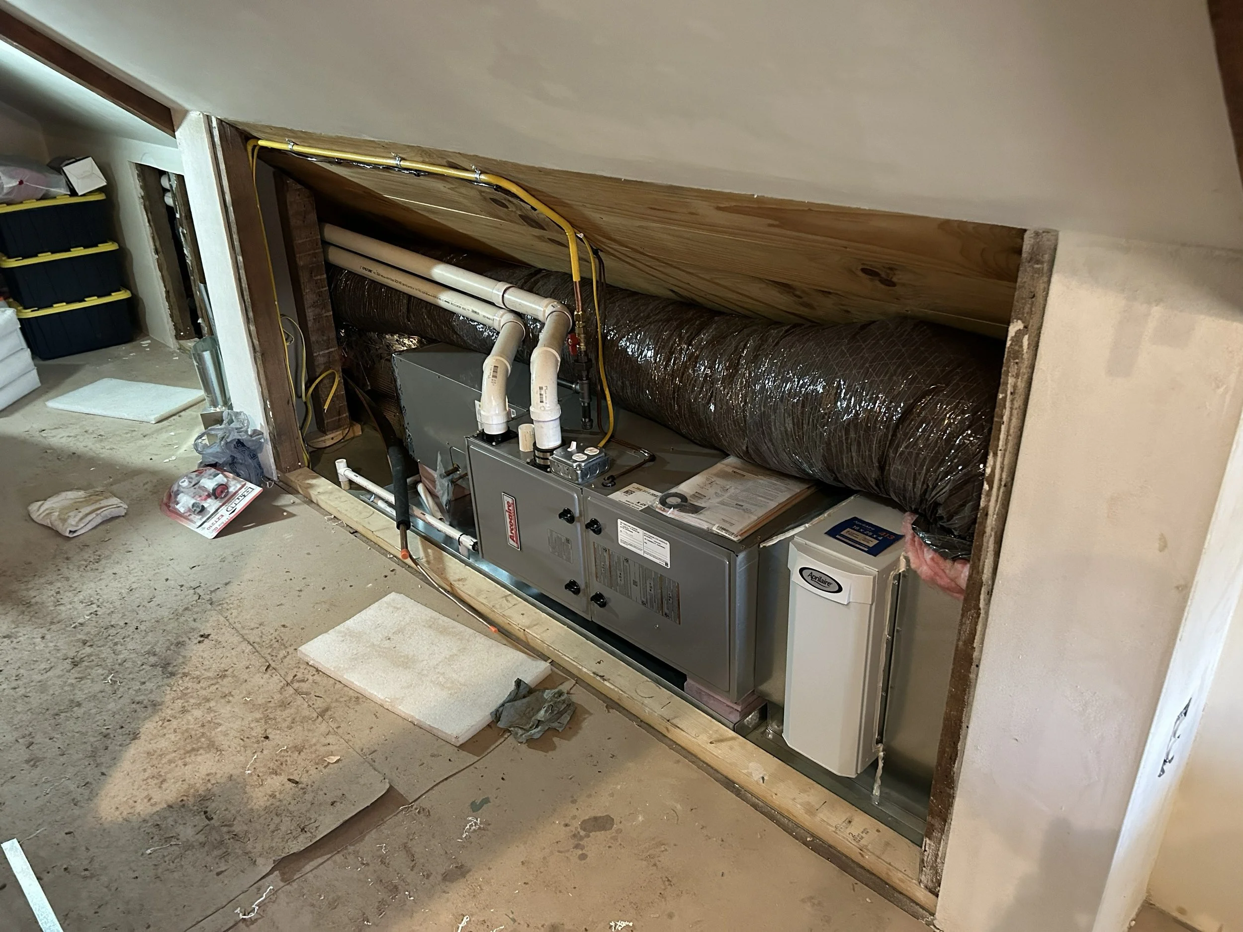 A residential furnace installation area with a furnace, ductwork, and piping on a concrete floor, partially boxed in with drywall and wooden framing.