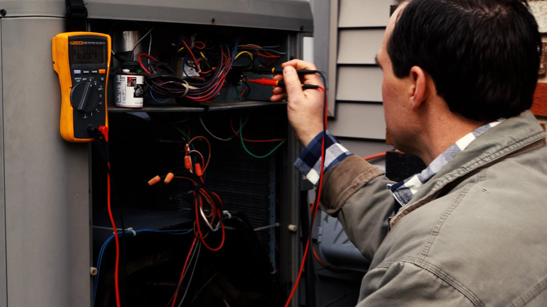 A man wearing a tan jacket and plaid shirt working on an electrical panel with multicolored wires, using a digital multimeter.
