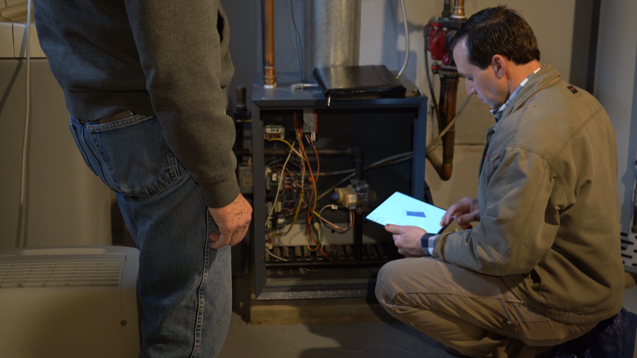 Two men working on an open electrical or mechanical device; one man is standing while the other is kneeling down and using a tablet.