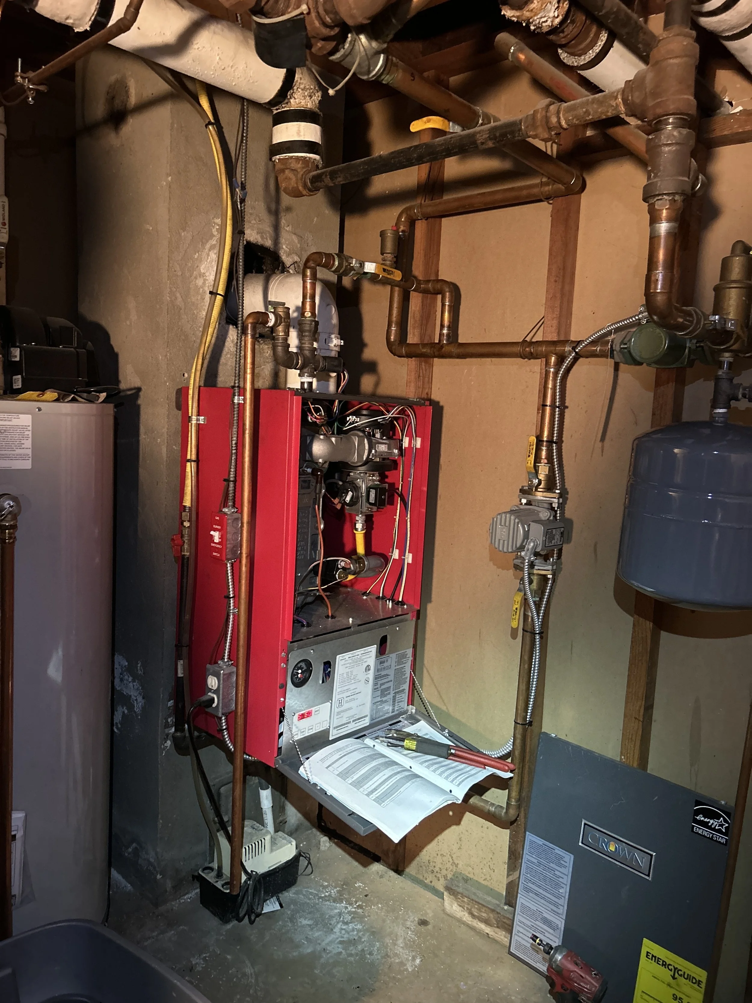 A utility room containing a red boiler with pipes, a water heater, and various plumbing components, with tools and an open instruction manual on the floor.