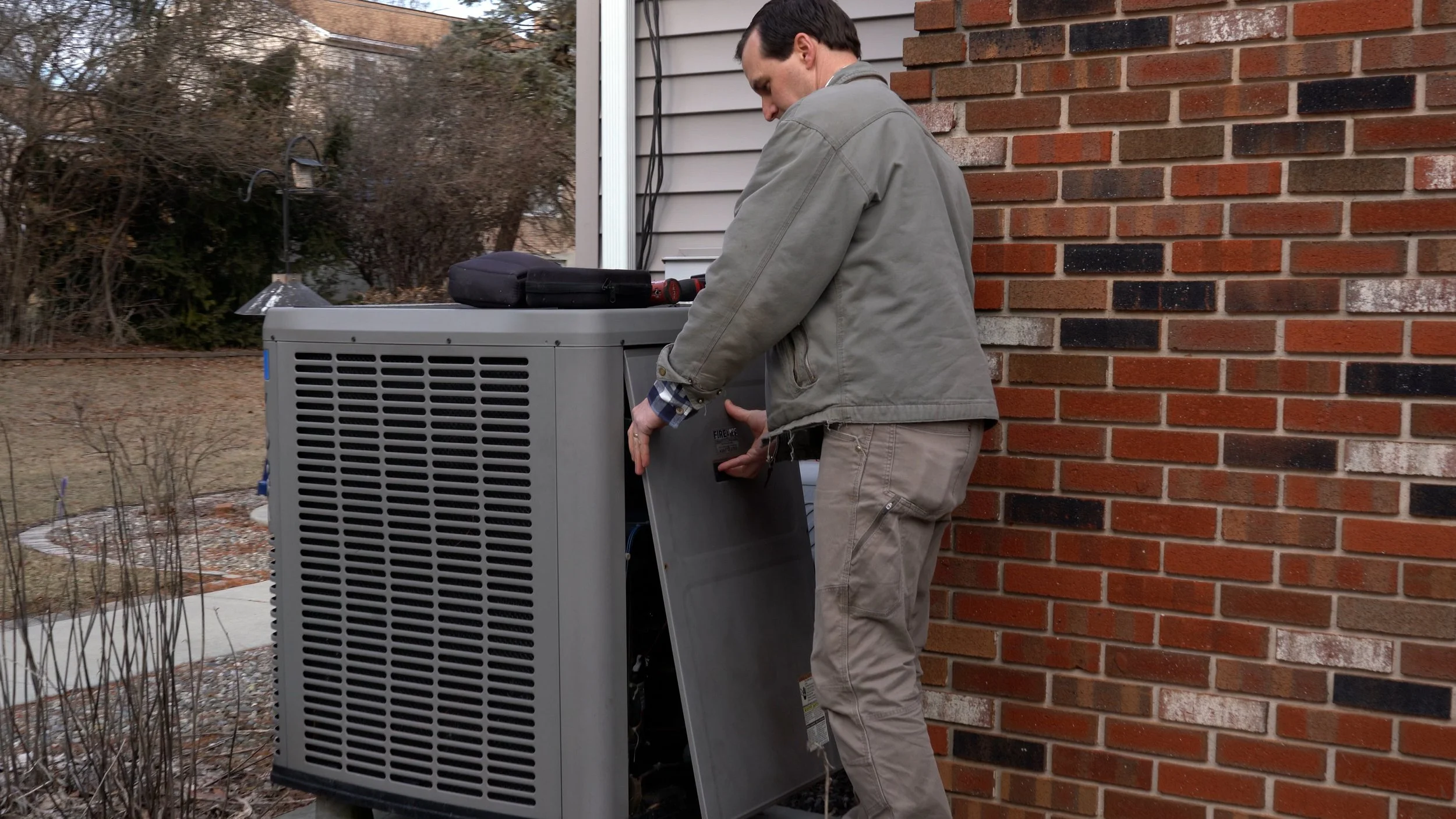 A man in a gray jacket and beige pants is repairing or inspecting an outdoor air conditioning unit next to a brick house wall, with tools on top of the unit and a toolbox nearby.