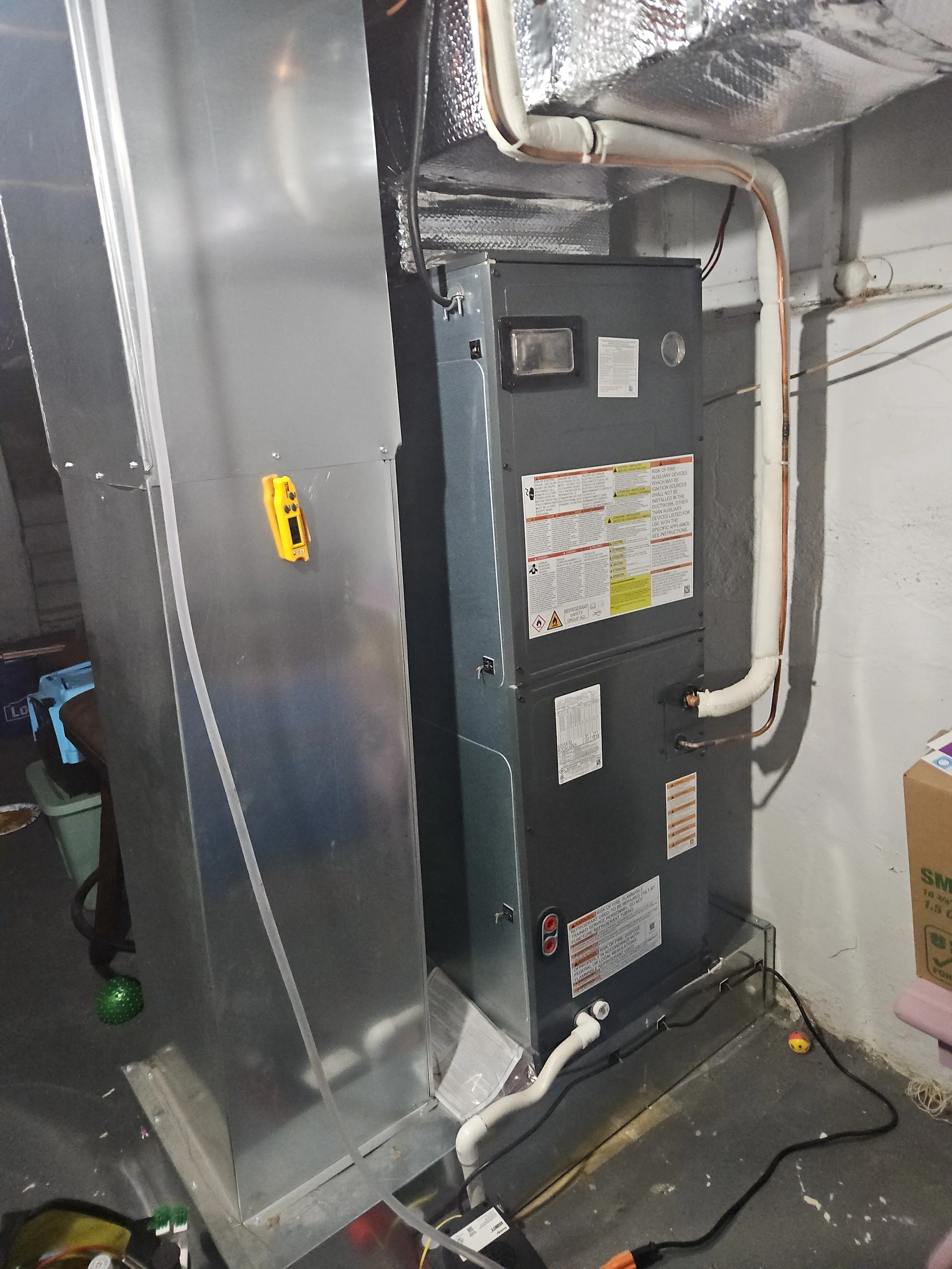 An HVAC furnace system installed in a basement with a silver metal duct, copper piping, and electrical connections. The furnace has labels and warning signs on its front panel.