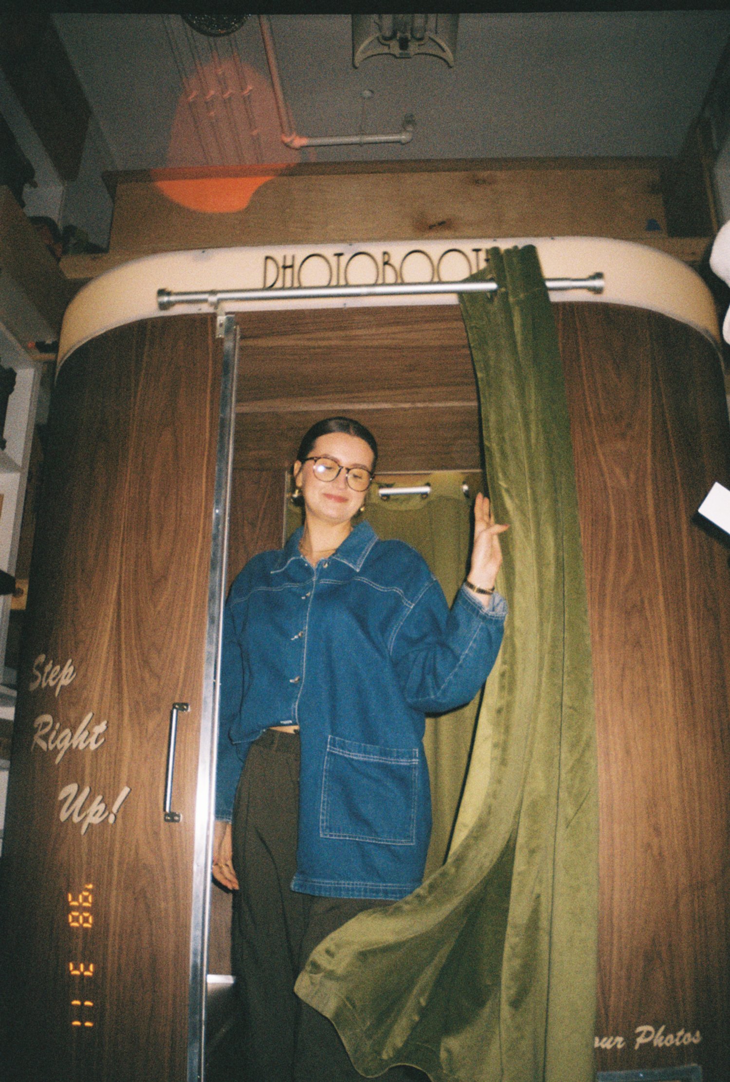A woman with glasses and dark hair standing inside a wooden photo booth with a green curtain. The booth has a sign that says 'Step Right Up!' and a label at the top that reads 'PHOTOBOOTH'.