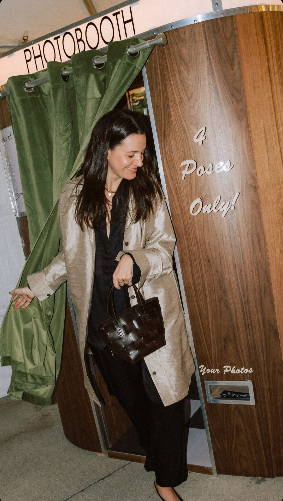 A woman with long dark hair, wearing a beige coat over black clothing, is smiling and looking down, holding a dark brown woven handbag as she steps out of a photo booth with a wooden exterior. The photo booth has the words '4 Poses Only!' written on 