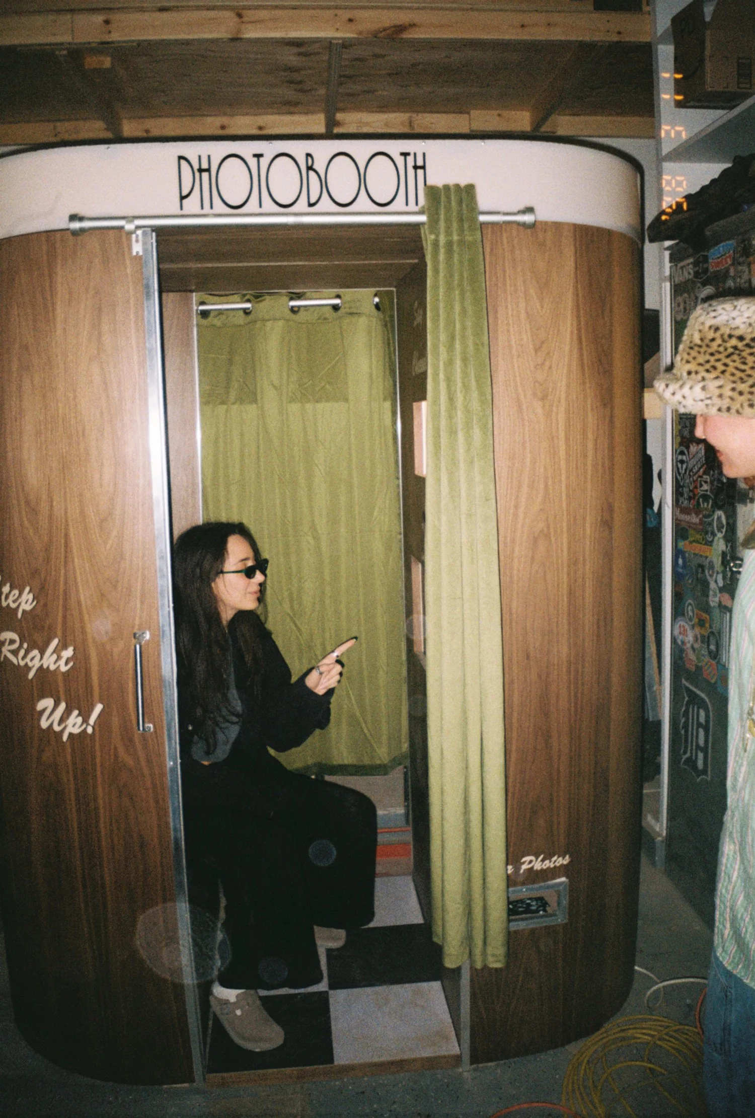 A woman sitting in a wooden photo booth, looking at her phone, with a yellow curtain inside. The booth has a sign that says 'PHOTOBOOTH' above it.