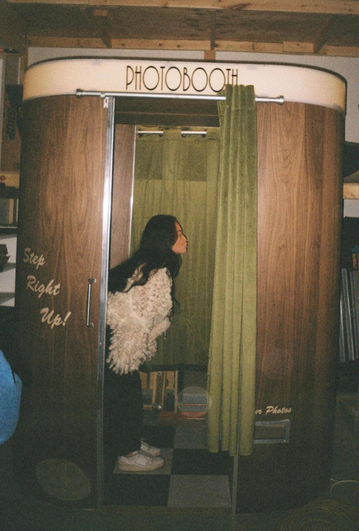 Woman with long dark hair wearing a white fluffy jacket and black pants inside a wooden photo booth with a green curtain, in a room with wooden ceiling beams and shelves.