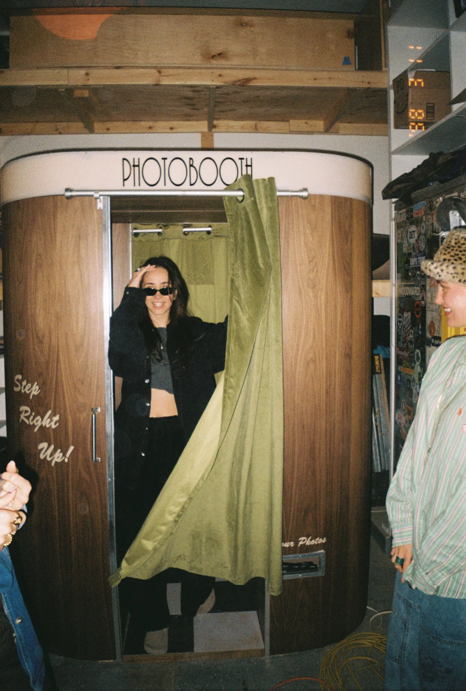 Woman wearing sunglasses and a black jacket standing inside a photo booth with a green curtain, smiling and waving. Another person is standing outside the booth, smiling and looking towards her. The photo booth has wooden sides and a white top with t