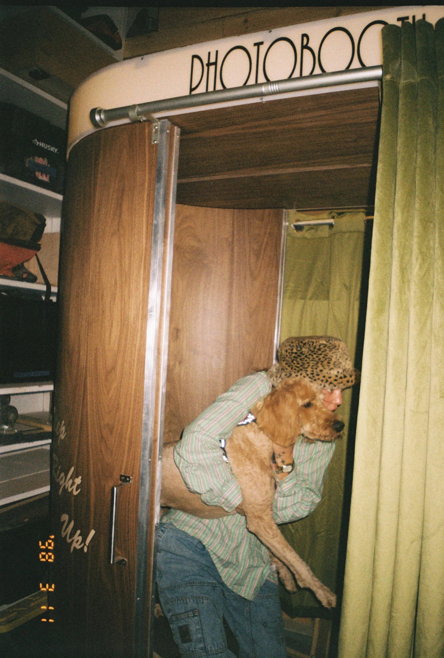 A person wearing a leopard-print hat and striped shirt holding a dog inside a photo booth with a wooden interior and a sign that reads 'PHOTO BOOTH' on top.
