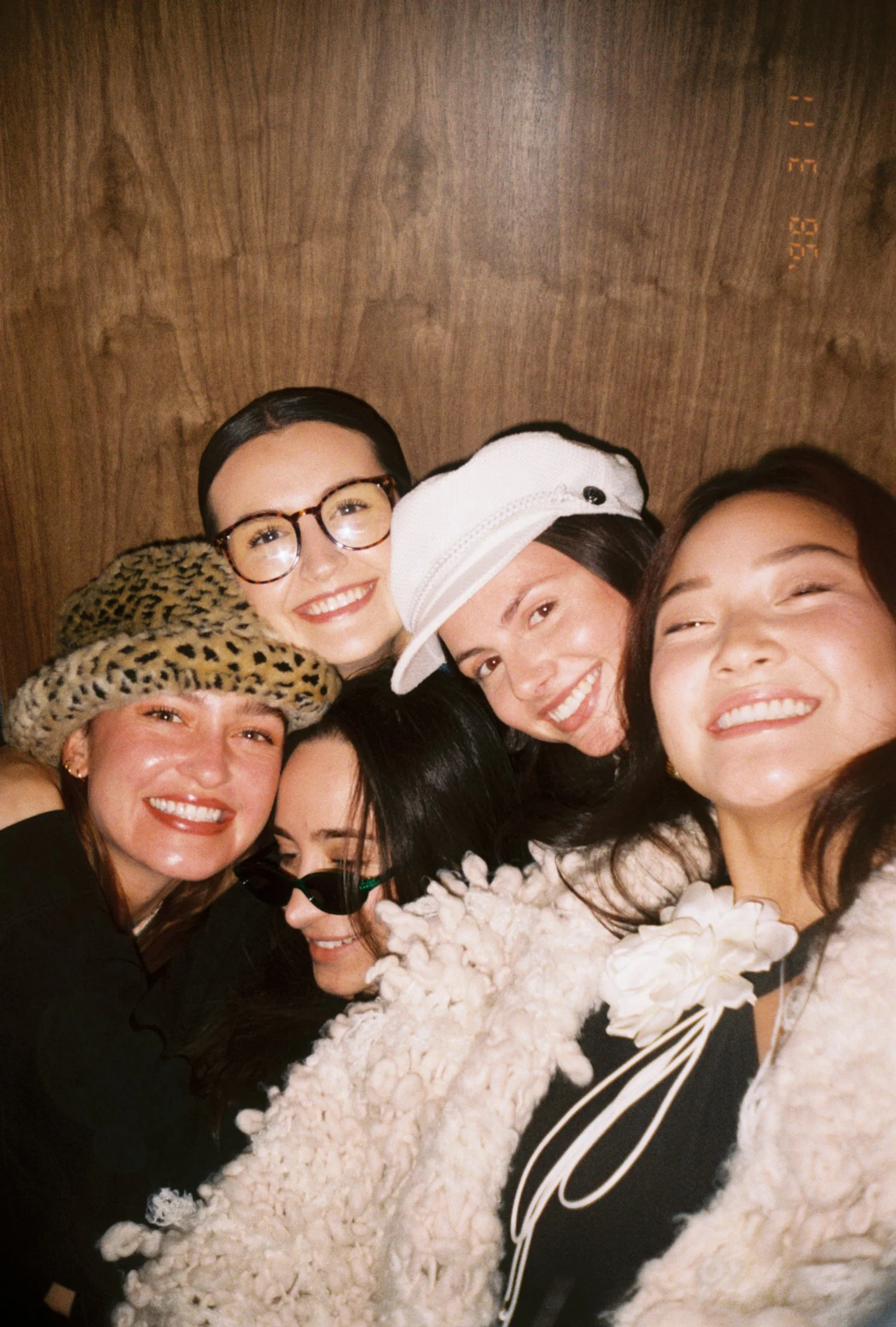 A group of five women smiling and posing closely together for a photo, wearing fashionable outfits and accessories, with a wooden background.